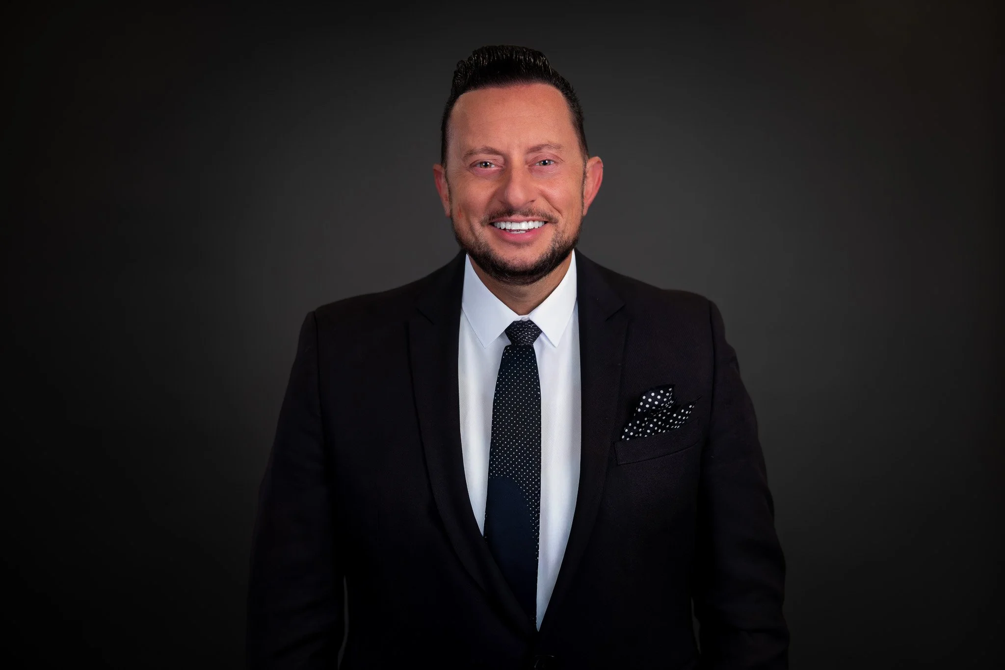 Portrait of a smiling man in a black suit, white shirt, and patterned tie, standing against a dark background.