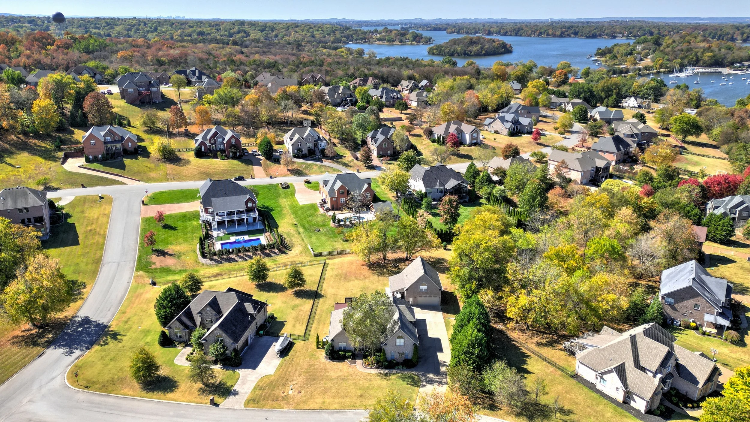 Aerial view of a suburban neighborhood with houses, green lawns, and trees, near a large lake with boats and islands, and a wooded landscape in the background.