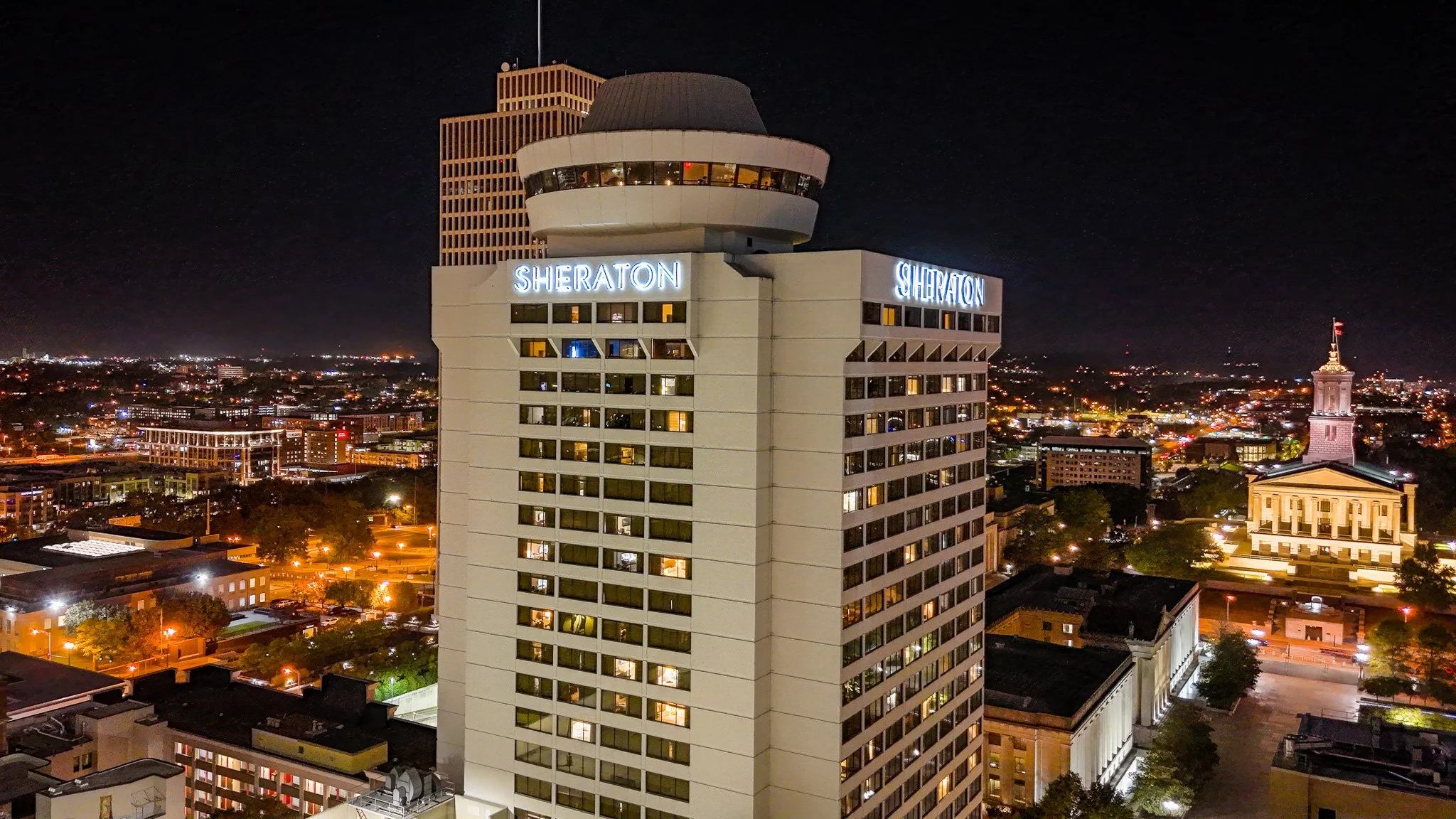 Nighttime cityscape with the Sheraton hotel tower illuminated and city lights in the background.