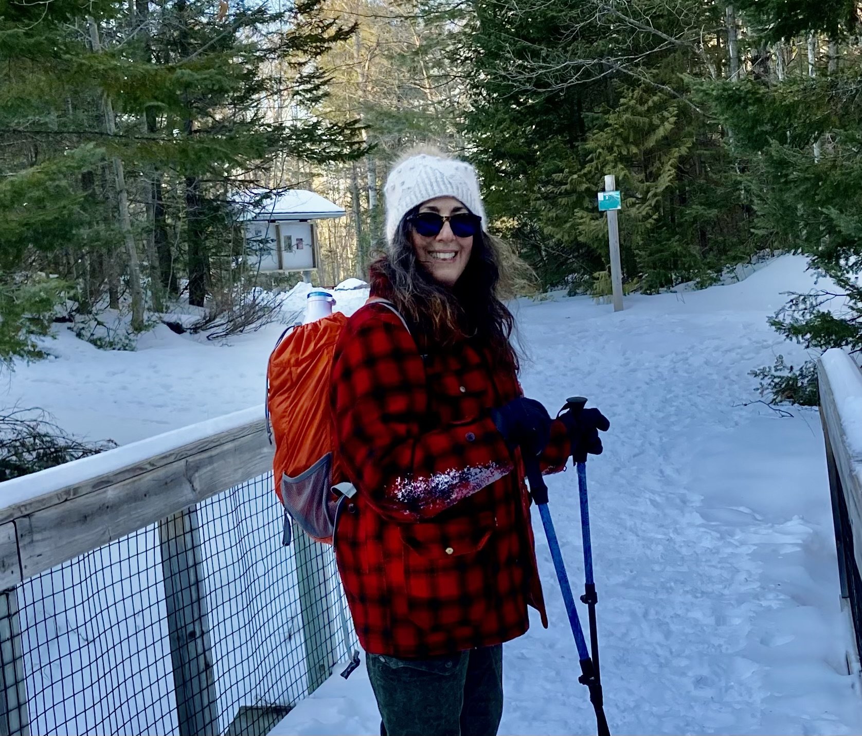 A woman with long curly dark hair and glasses smiling outdoors with green leafy background.