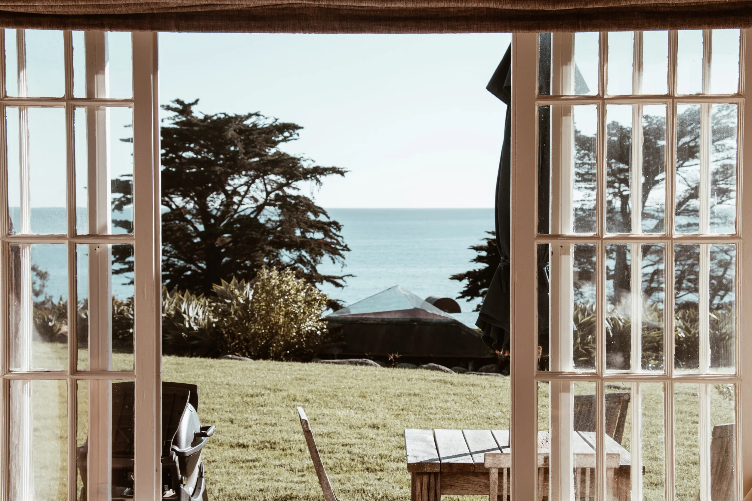 View of a backyard through glass-paneled French doors, showing a lawn, outdoor furniture, trees, and the ocean in the distance.