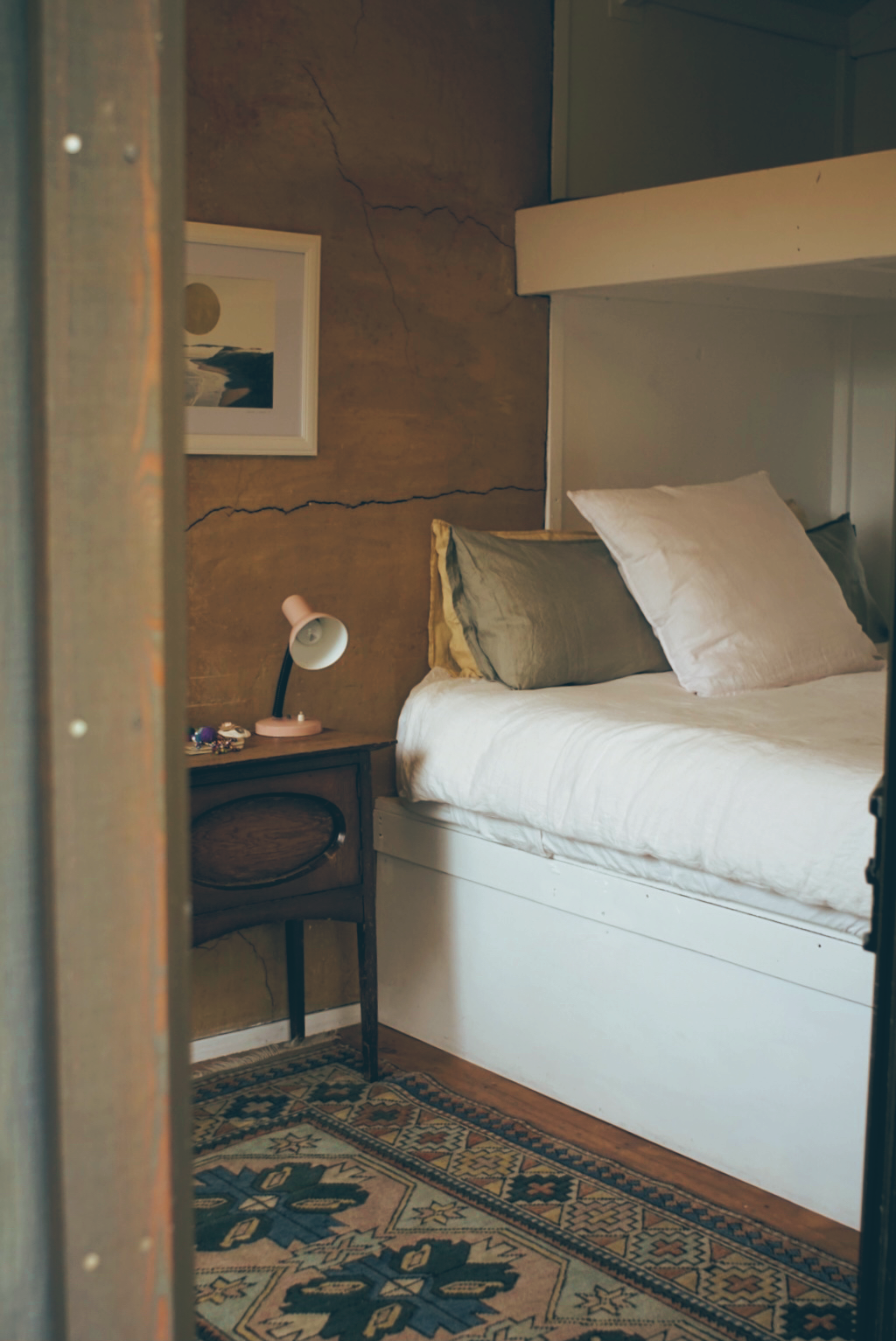A cozy bedroom with a bed, pillows, a small wooden nightstand with a pink lamp, a framed picture on the wall, and a patterned rug on the floor.