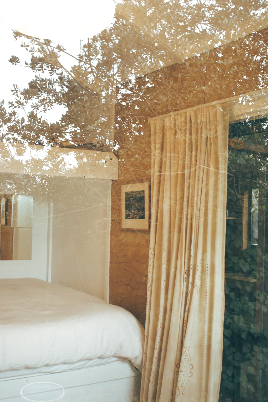 Reflection of a bedroom with yellow curtains, framed picture, and outside trees seen through a window.