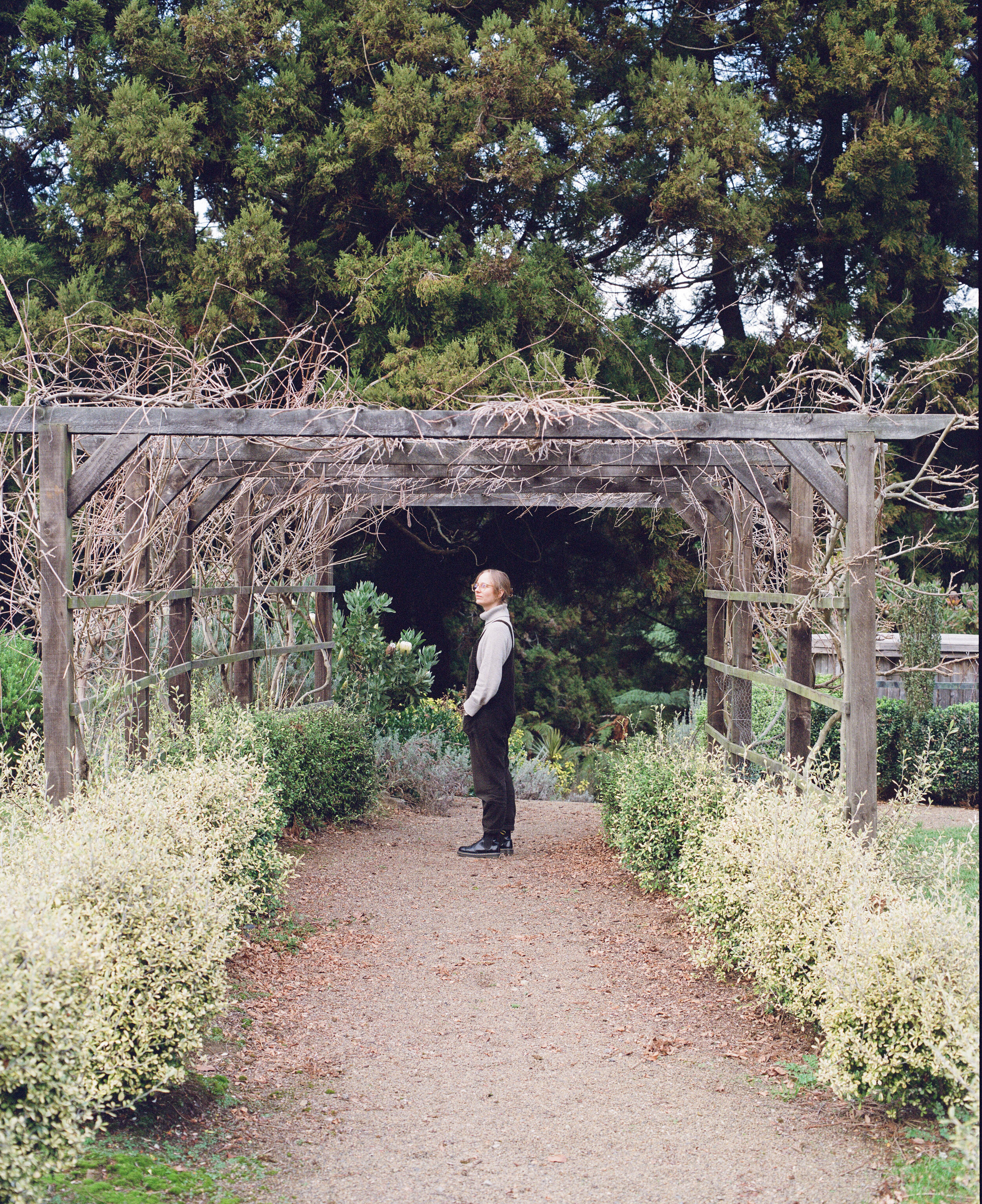 A woman with glasses and a gray sweater standing on a gravel path in a garden with a trellis overhead and green bushes on both sides.