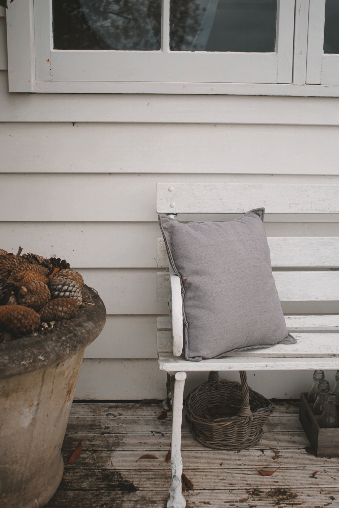 A white wooden bench with a gray pillow on it, next to a large terracotta pot filled with pinecones, on a weathered wooden porch with white siding and a window.