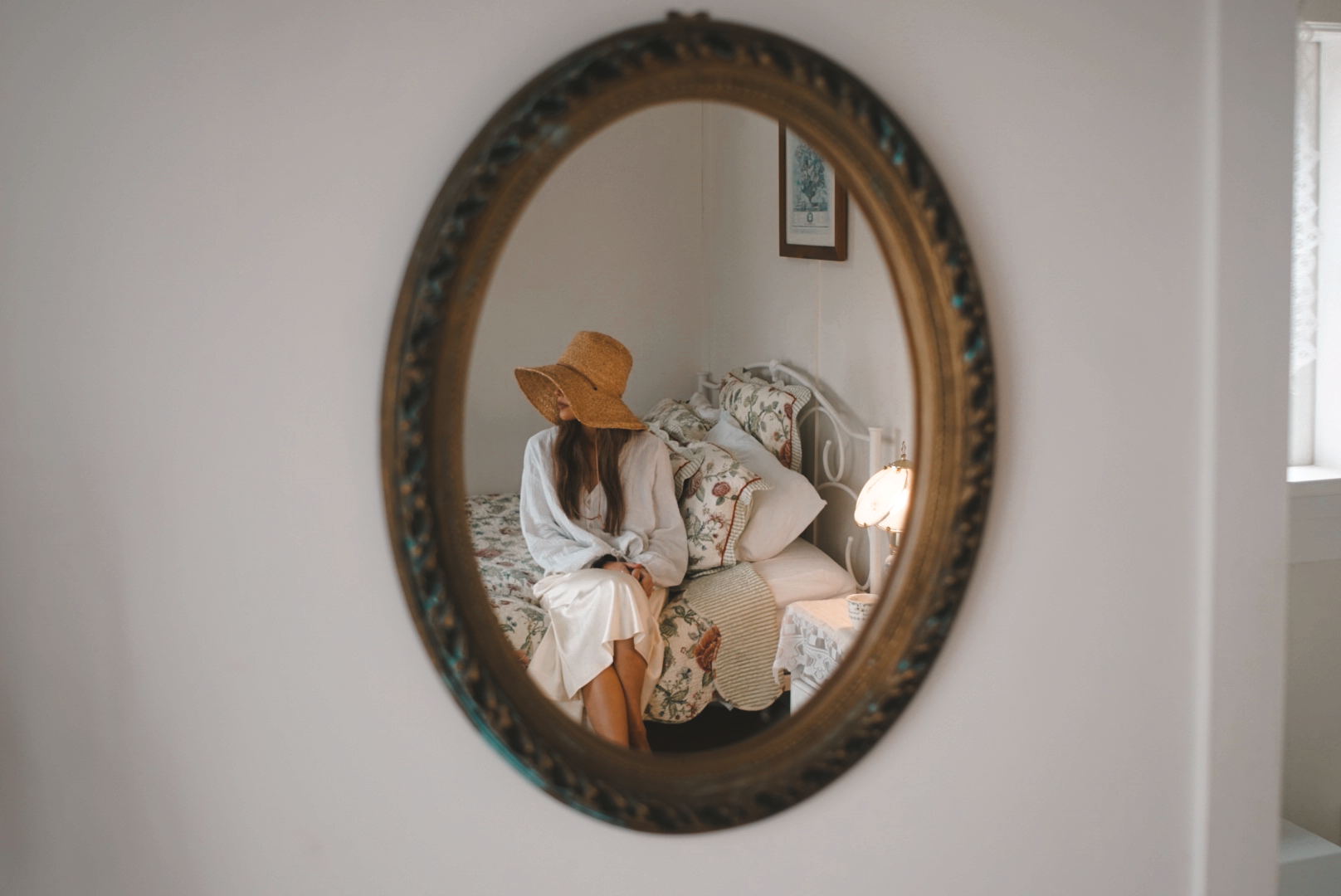 A woman sitting on a bed, wearing a large straw hat and white dress, is viewed through a round mirror in a bedroom with floral bedding and a small lamp.