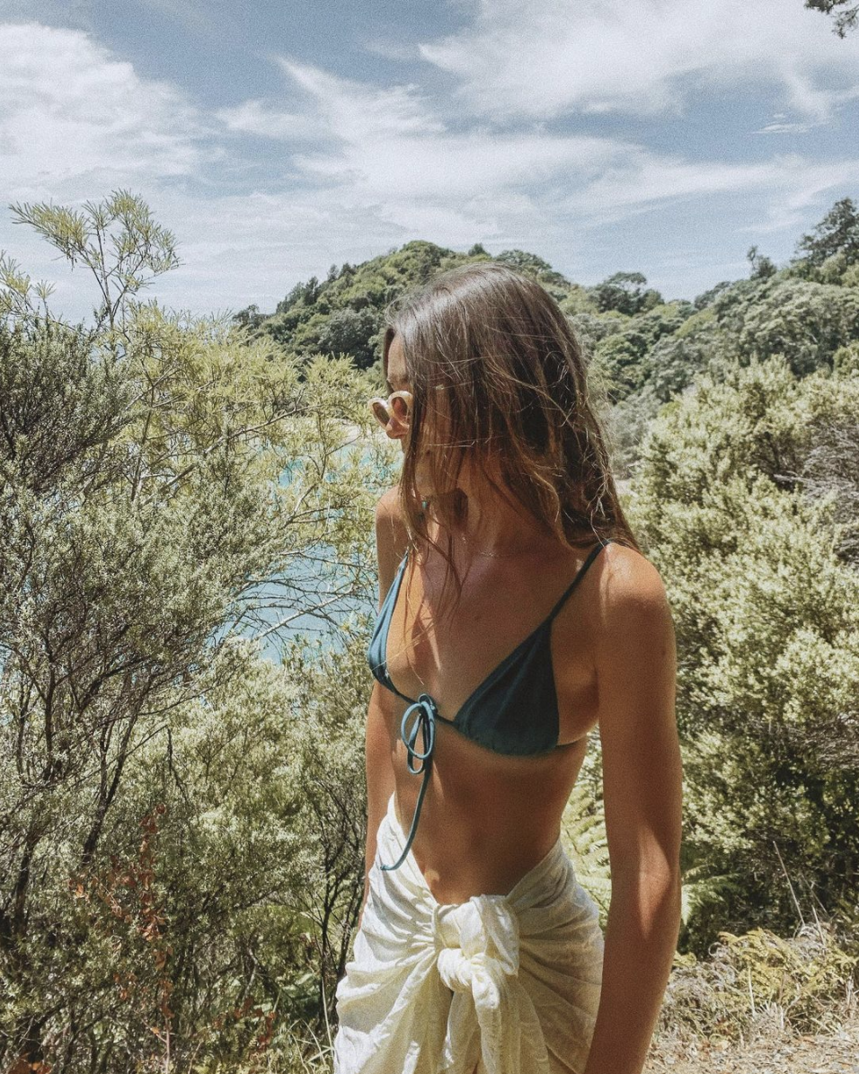 A woman in a blue bikini top and sunglasses stands outdoors among green shrubs and trees, with a scenic landscape and cloudy sky in the background.
