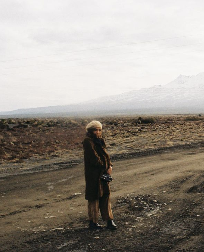 Woman dressed in warm clothing, holding a camera, standing on a dirt road in a barren landscape with snow-capped mountains in the distance.