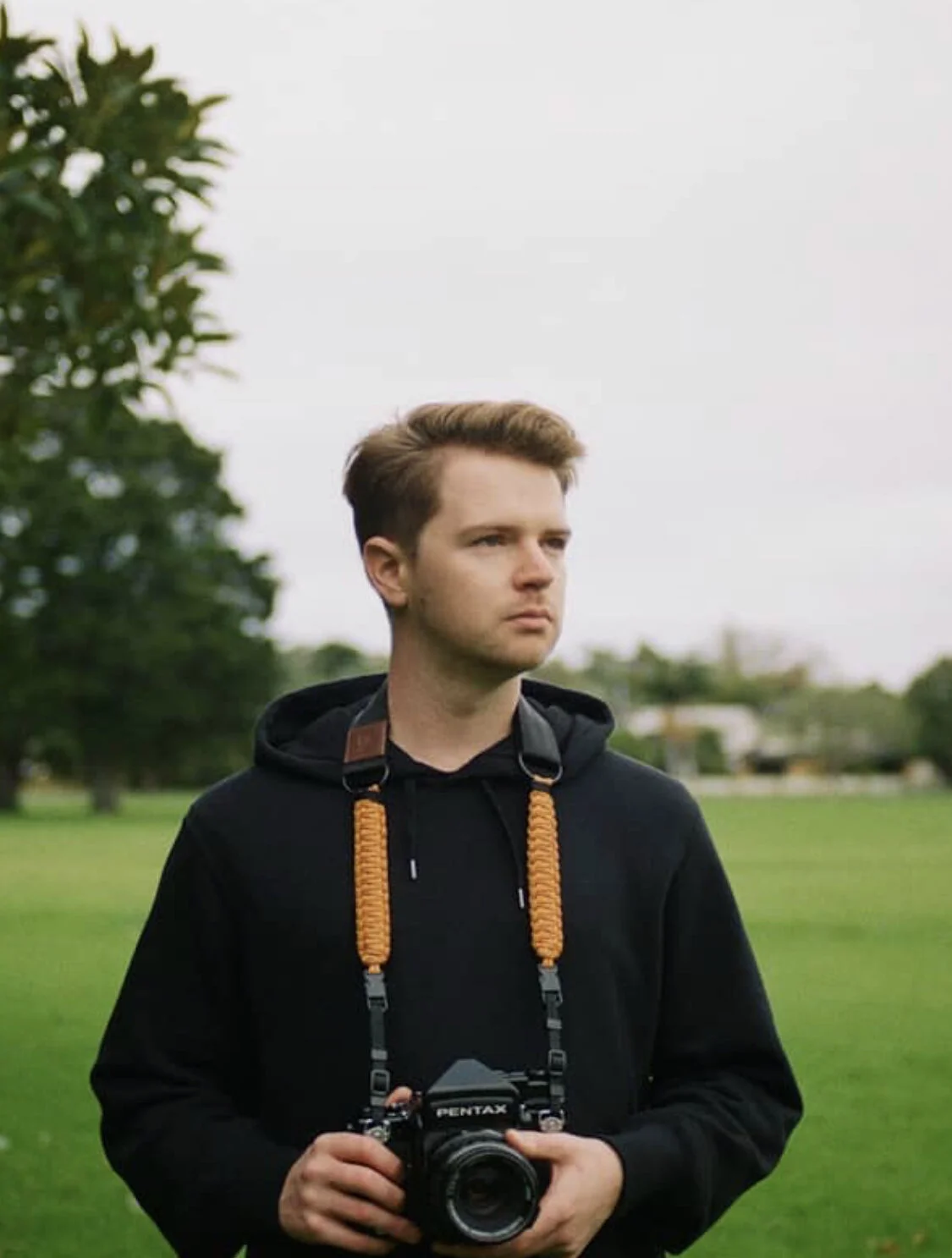 Young man standing outdoors on a cloudy day, holding a Pentax camera, wearing a black hoodie with a camera strap around his neck, with green trees and grass in the background.