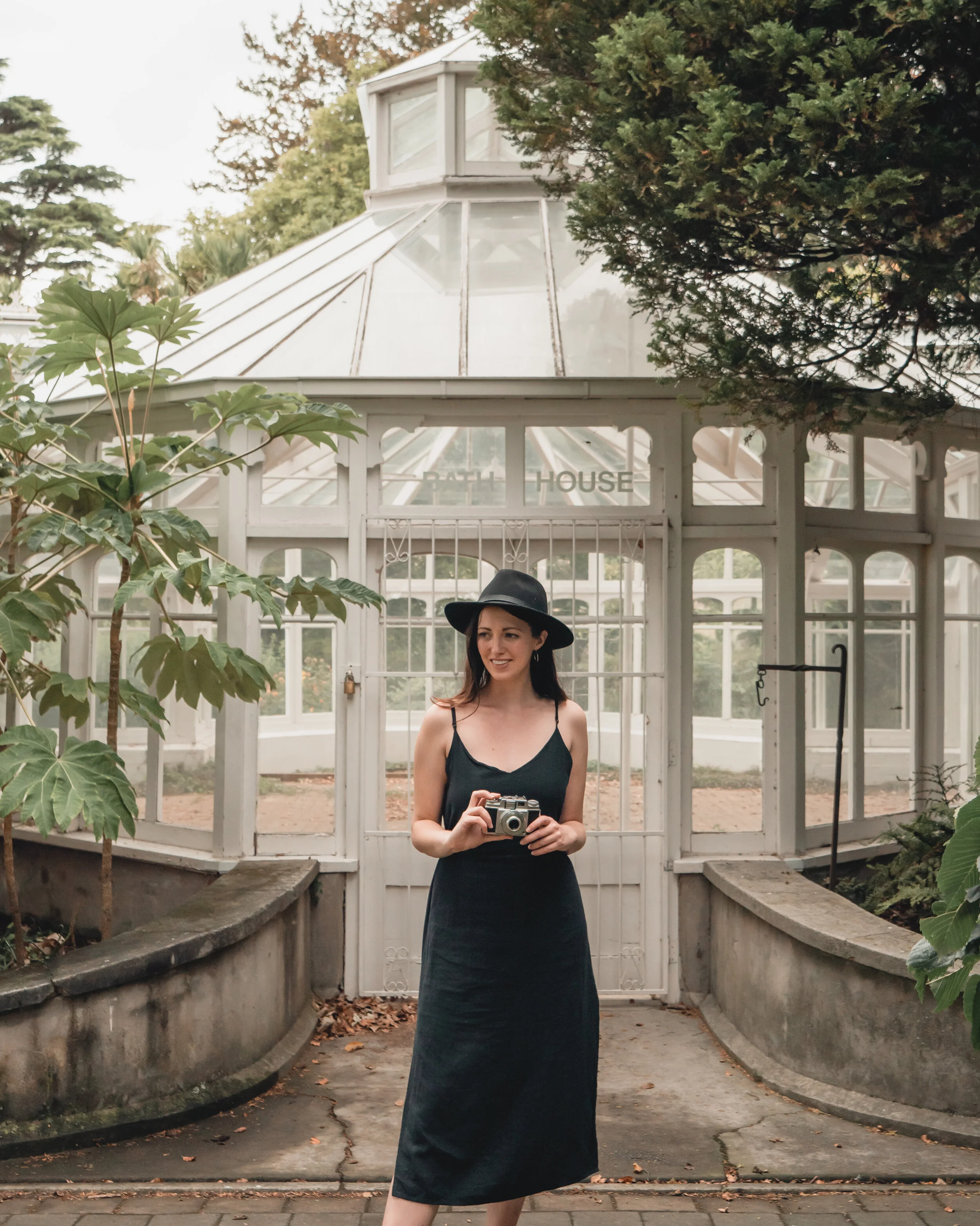 A woman standing outdoors in front of a white ornate greenhouse with large glass windows, holding a camera, smiling, wearing a black dress and black hat, surrounded by green trees and plants.