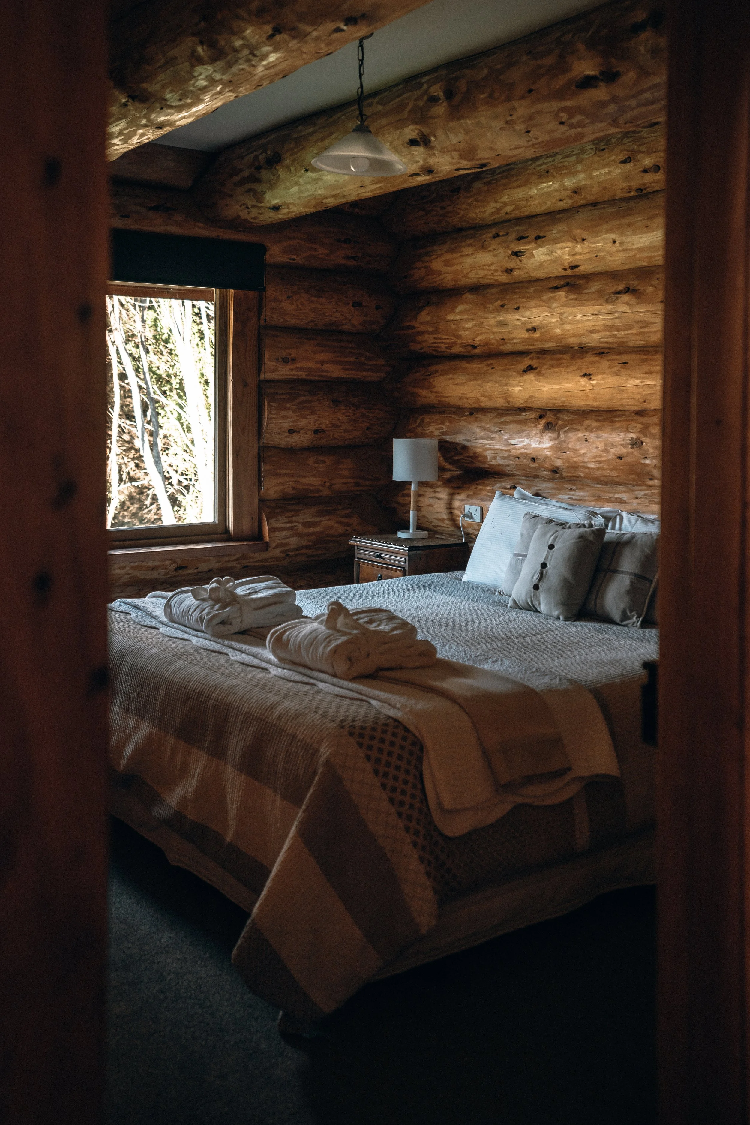 A cozy bedroom with wooden log walls and a window showing a view of trees outside. The bed is made with a beige and brown checkered quilt, multiple pillows, and has towel swans placed on it. There is a bedside table with a white lamp and some electronic outlets.