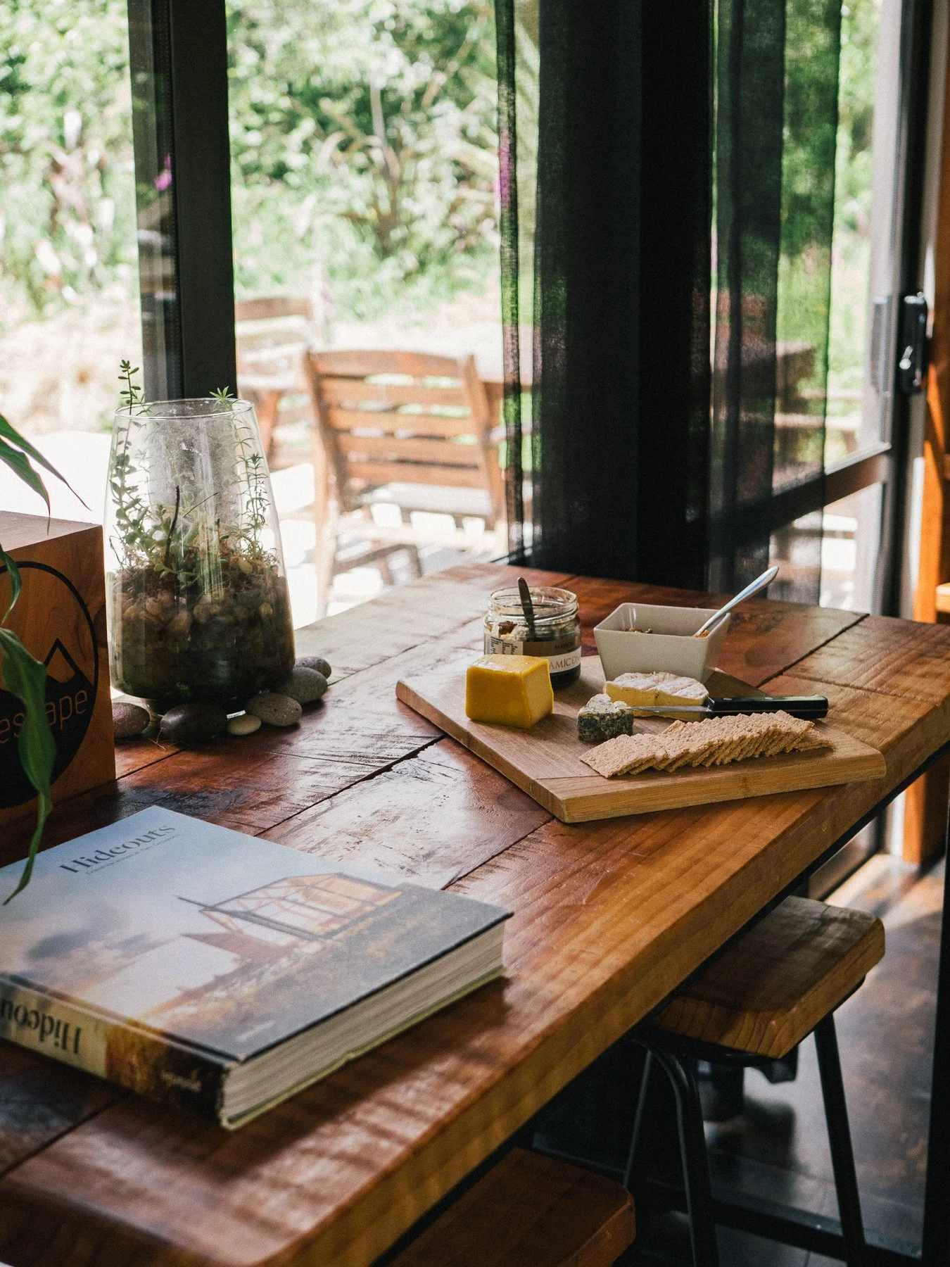 A wooden table near a window with black curtains, holding a glass terrarium with plants, a jar of jam, a cheese wedge, a small dish with a spoon, some crackers, and a book titled 'Hidcots' with a house illustration.