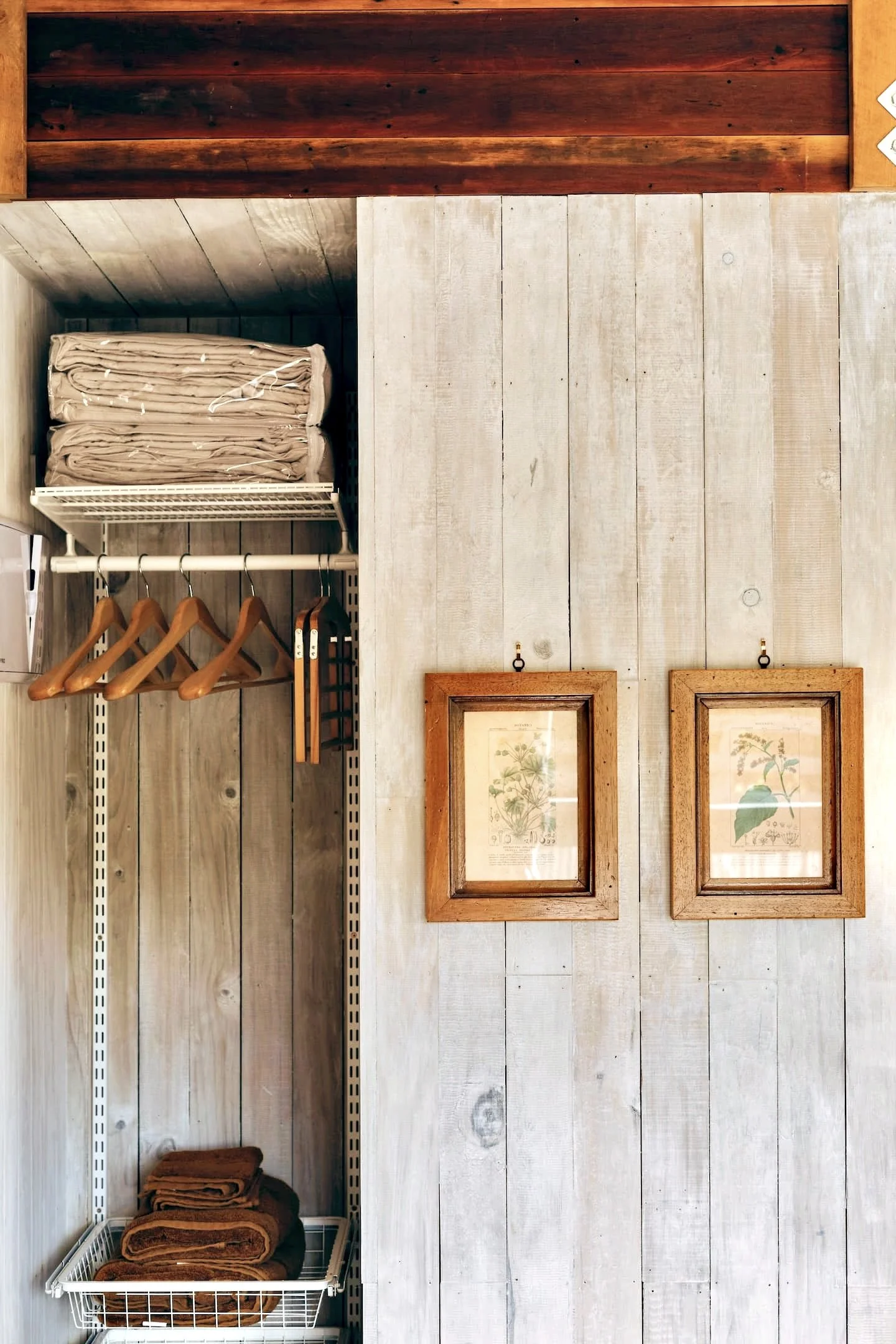 Wooden wall with framed botanical prints, open closet with hanging brown towels, and a shelf with folded beige linens.