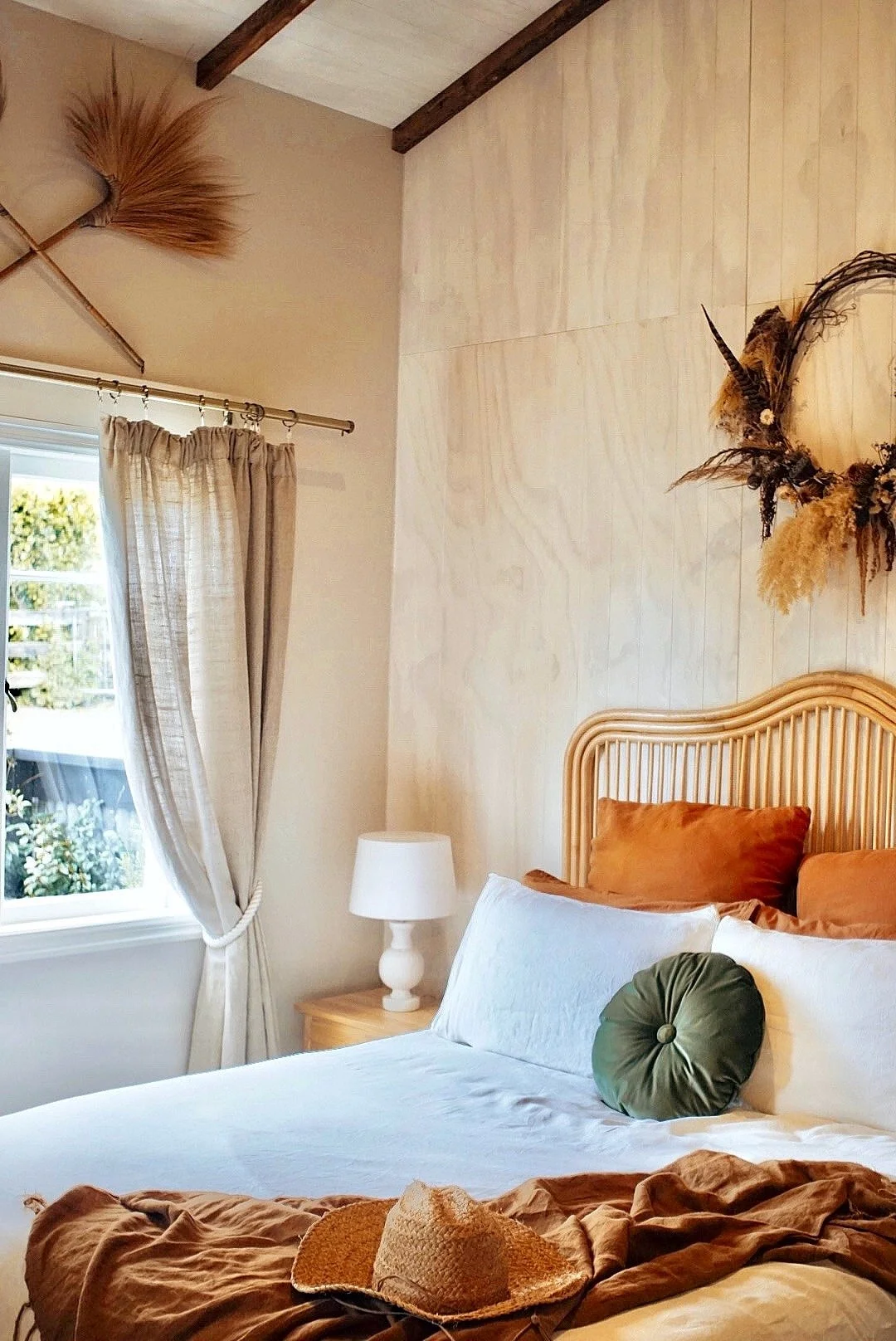 A cozy bedroom with a bed featuring white sheets, a green round decorative pillow, and rust-colored pillows against a wooden headboard. Next to the bed, there is a wooden nightstand with a white lamp. A window with beige curtains offers a view of greenery outside. The wall behind the bed is decorated with a dried floral wreath, and a broom made of natural materials is hung on the upper left corner of the wall.