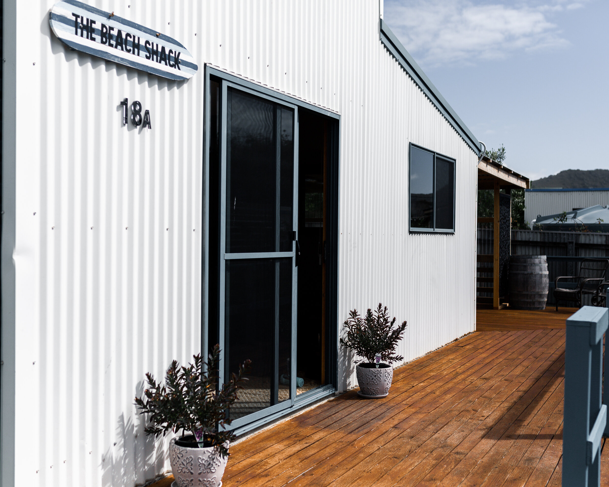 Exterior of a beachside building with a sign that says 'The Beach Shack,' a sliding glass door, and a window. There are two potted plants, a wooden deck, and outdoor seating with mountains in the background.