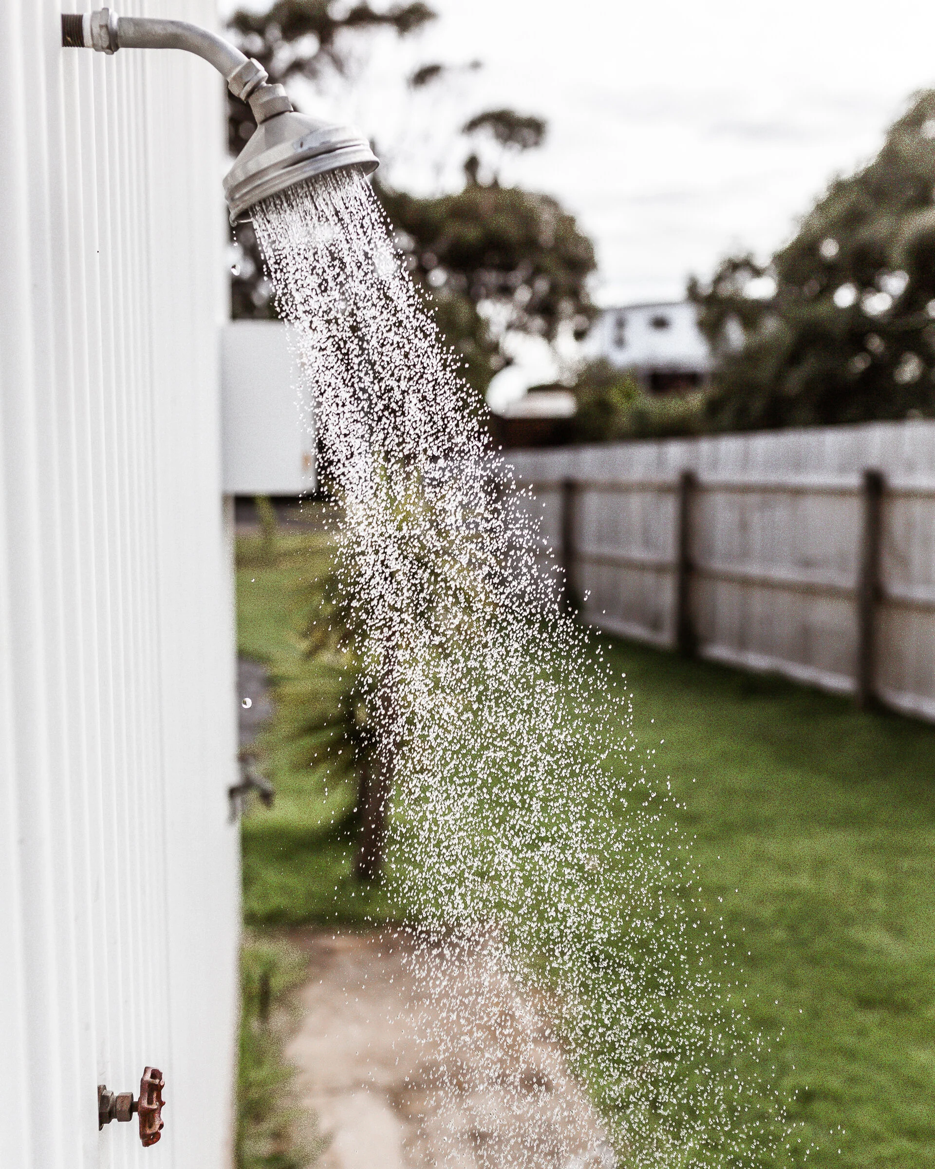 Close-up of outdoor shower with water spray, backyard with fence and trees in background
