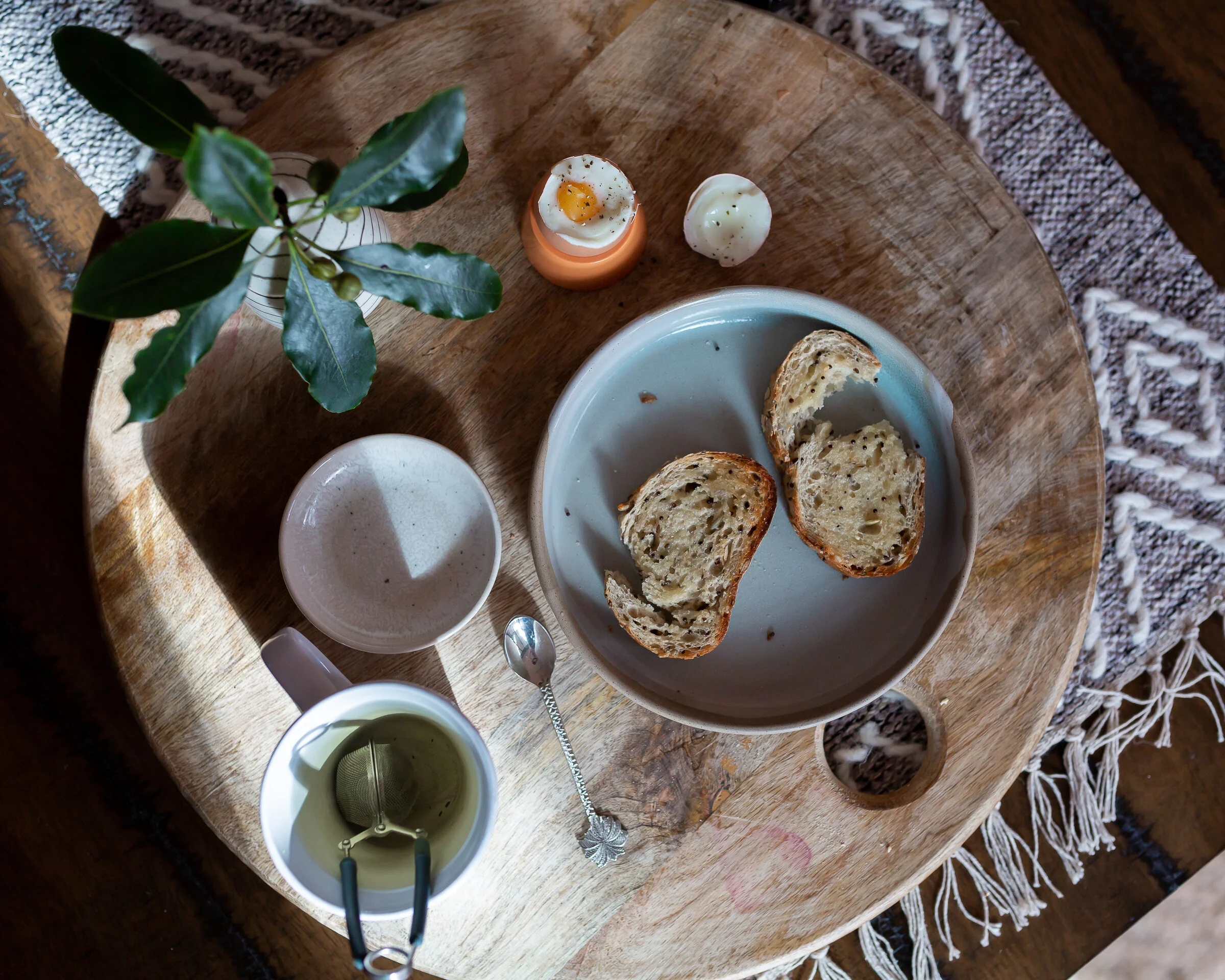 A round wooden tray with a small plant, a soft boiled egg in an egg cup, a hard boiled egg, a white bowl, a white cup with tea, a spoon, and a plate with two slices of bread on it.