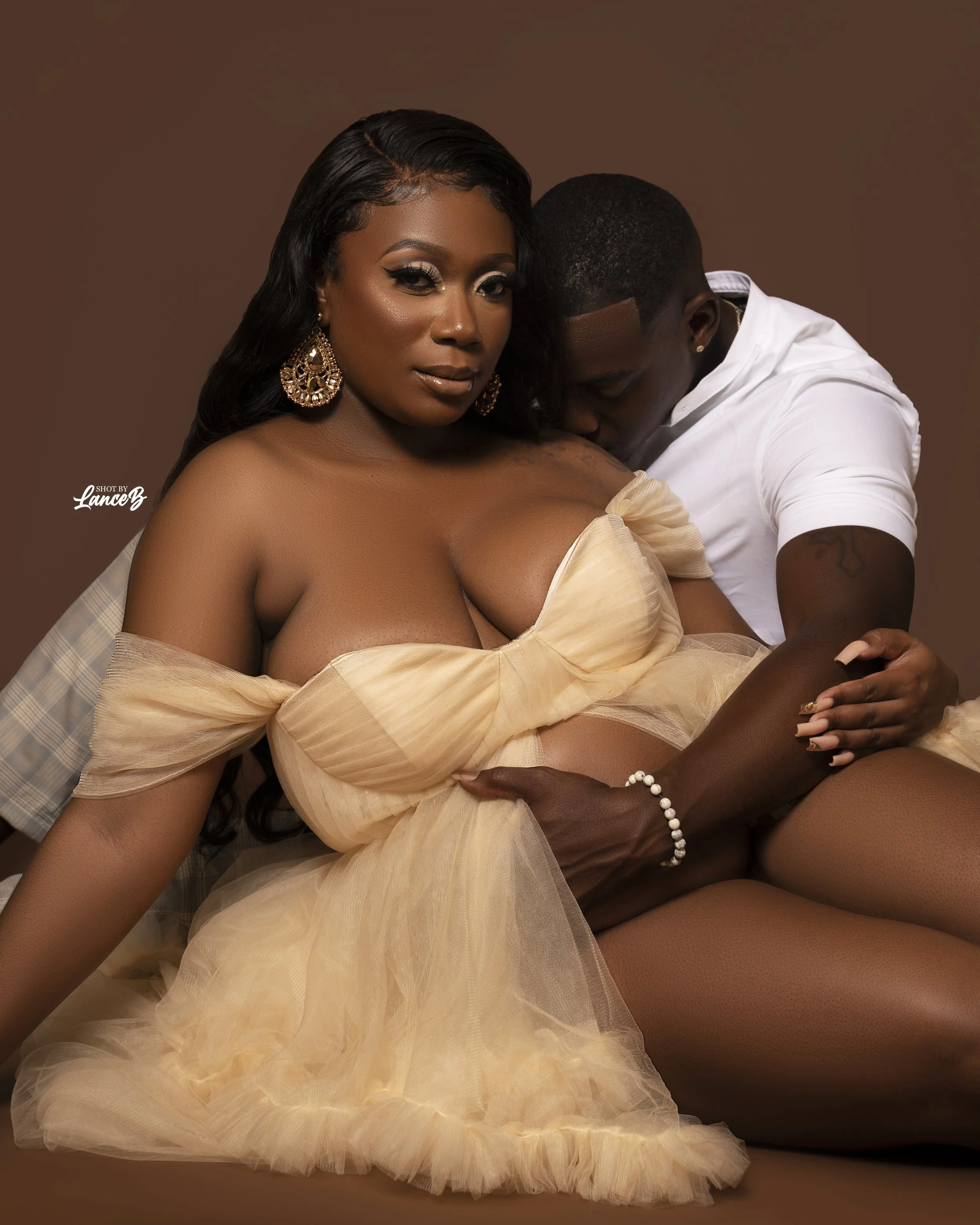 A woman with dark hair and large earrings sits on a man's lap, wearing a beige off-the-shoulder tulle dress. The man, dressed in a white shirt, leans in close, his face near her shoulder, and holds her arm. They are posed against a plain brown background.