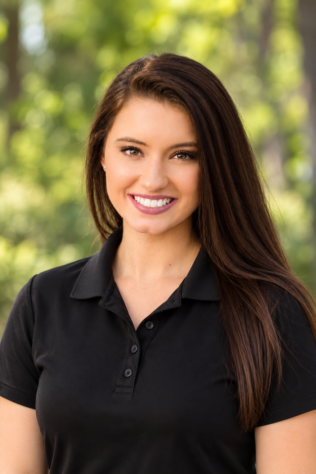 A young woman with long brown hair and a bright smile, wearing a black polo shirt, outdoors with blurred green trees in the background.