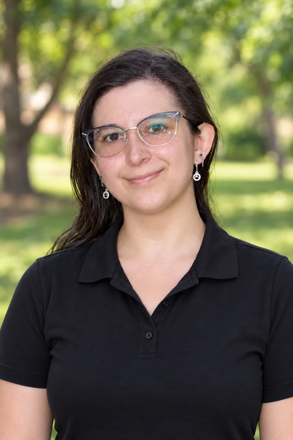 A young woman with dark hair, glasses, and earrings, wearing a black collared shirt, standing outdoors with trees and greenery in the background.