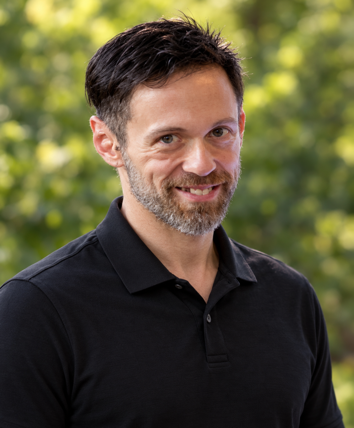 A man with short dark hair, a beard, and a mustache, wearing a black collared shirt, smiling outdoors with blurred green trees in the background.