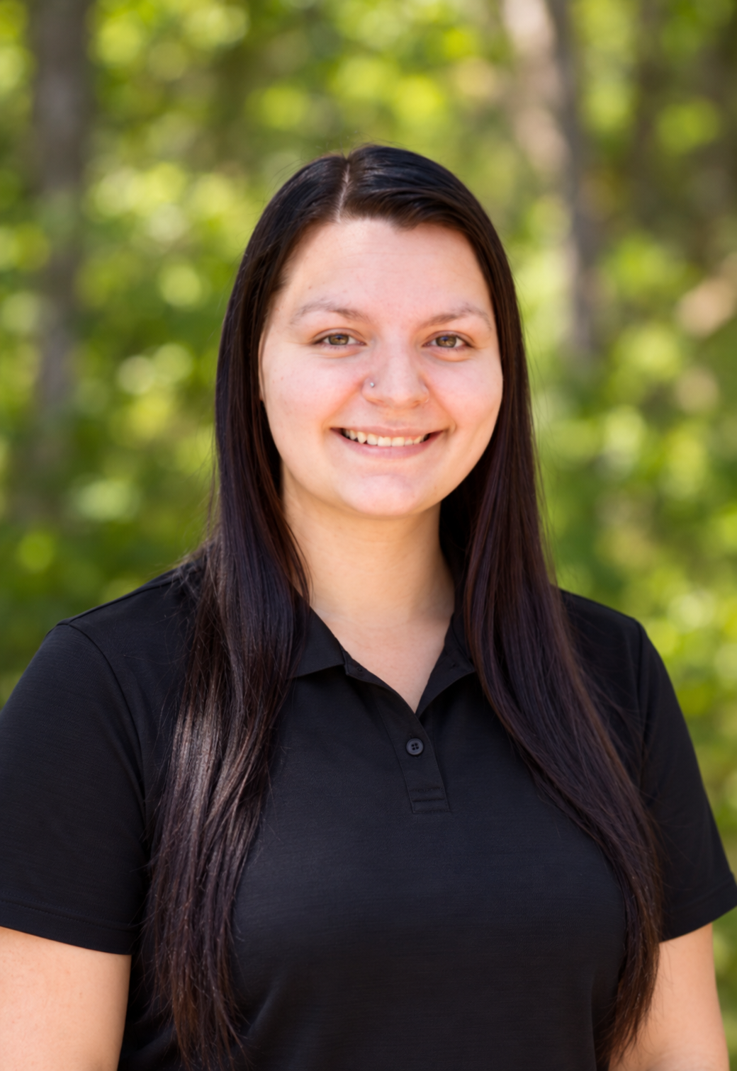 A young woman with long dark hair and light skin smiling outdoors with a background of green trees, wearing a black collared shirt.