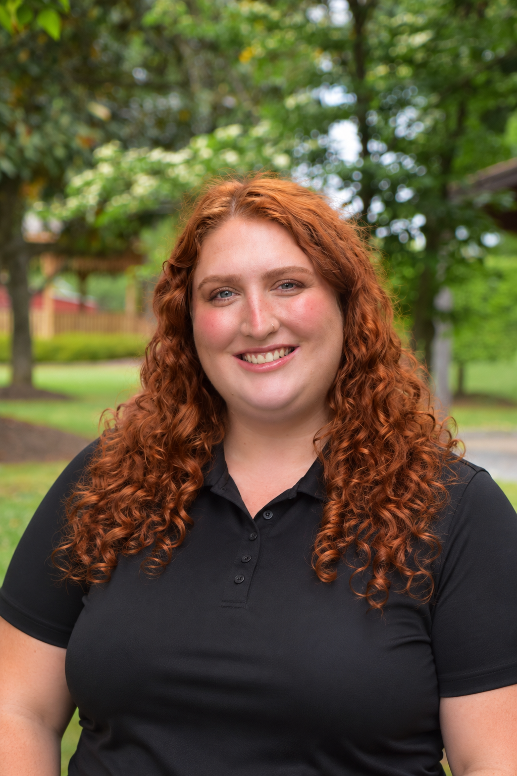 Headshot of a woman with long, curly red hair, wearing a black polo shirt, outdoors with trees and greenery in the background, smiling.