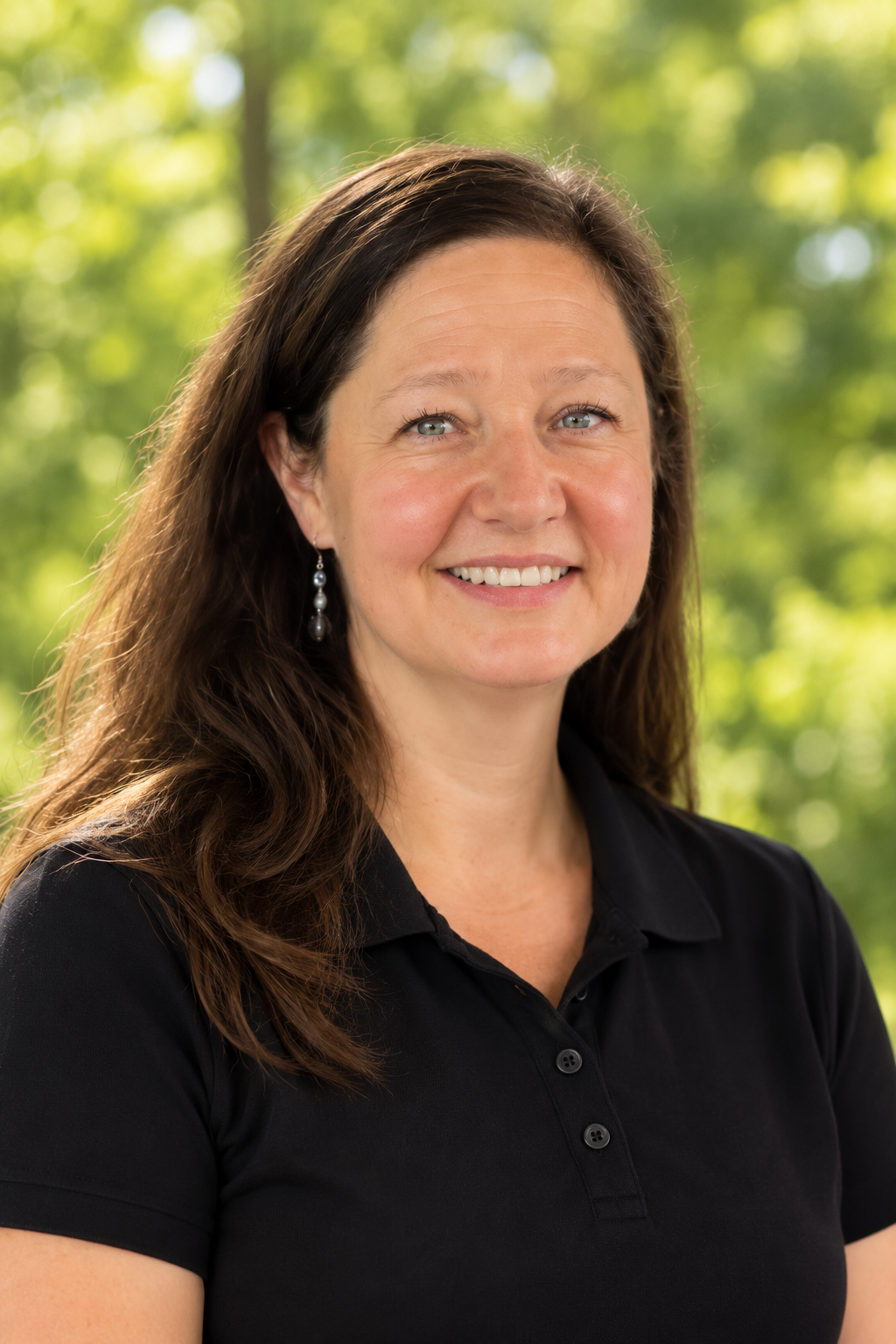 A woman with long, dark brown hair, smiling, wearing a black polo shirt and pearl earrings, with a blurred green, leafy background.