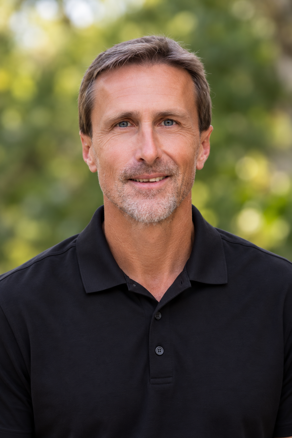 A middle-aged man with short brown hair, a beard, and blue eyes, wearing a black polo shirt, smiling outdoors with blurred green trees in the background.