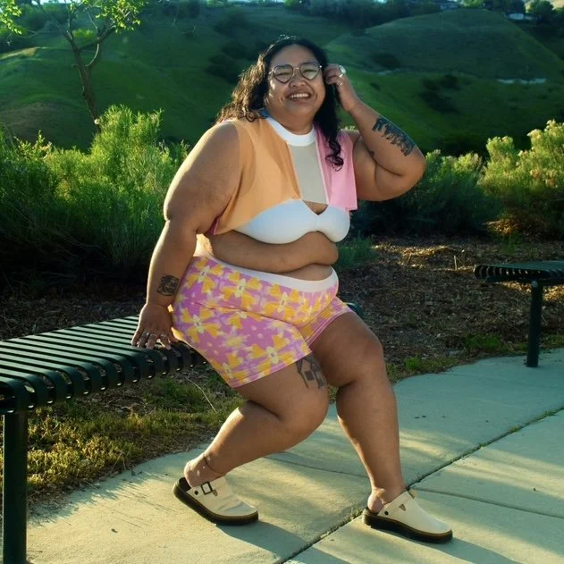 A smiling person with glasses is sitting on a park bench, wearing colorful shorts and a sleeveless top, with greenery and hills in the background.