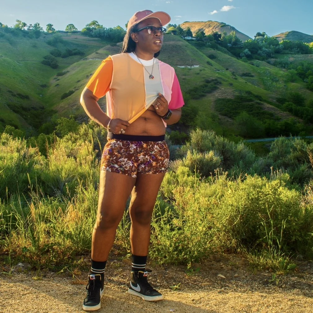 A person standing outdoors in a hilly, green landscape during sunset, wearing sunglasses, a pastel-colored cropped jacket, a white t-shirt, floral shorts, socks, and sneakers.