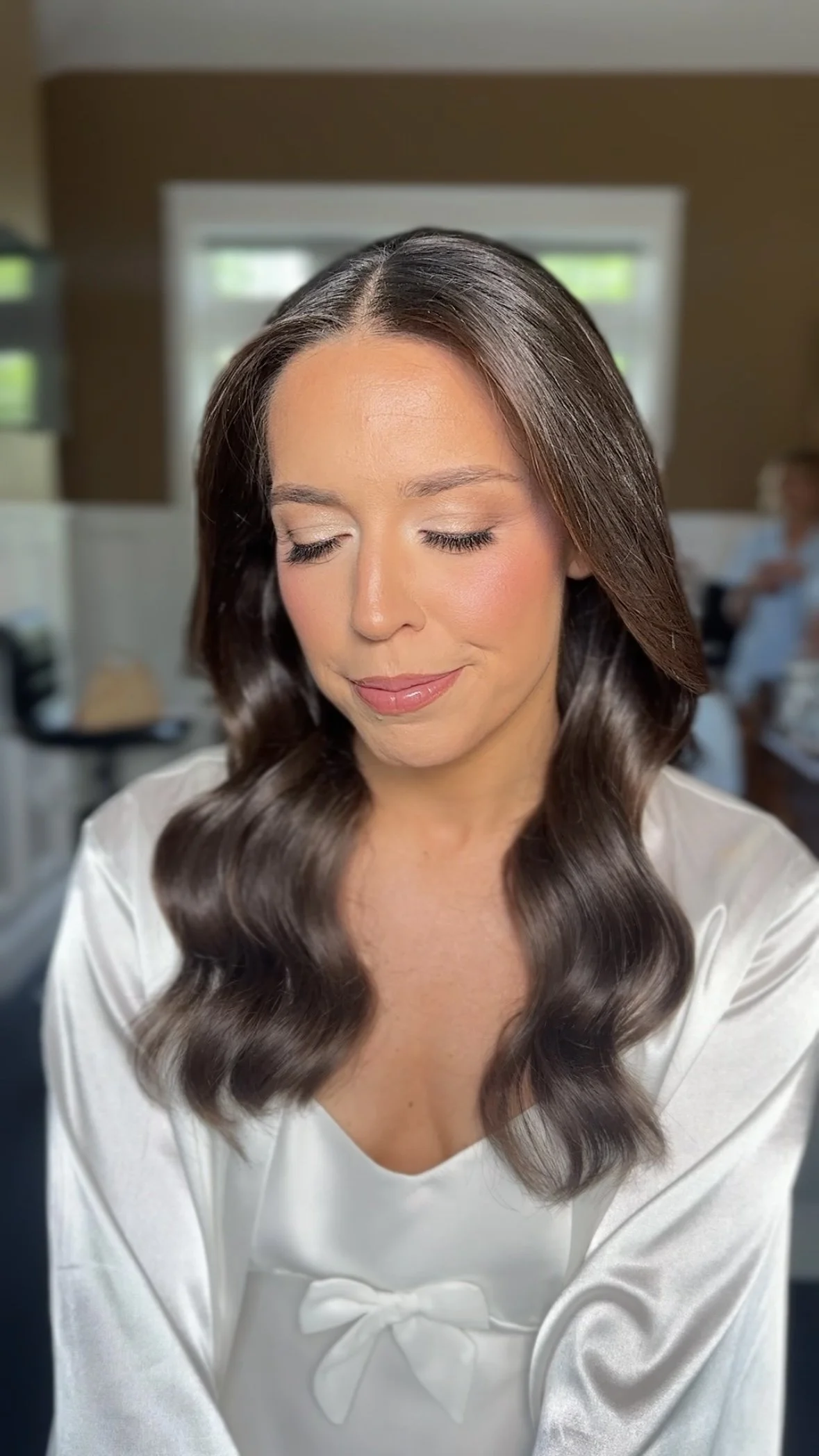 A woman with long, wavy brown hair, wearing a white satin blouse with a bow, smiling softly with eyes closed in a cozy indoor setting.