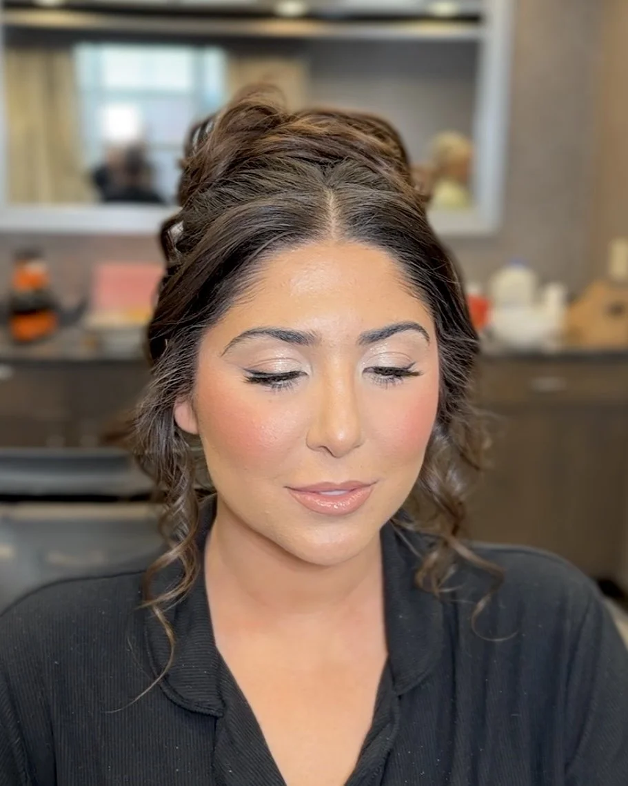 A woman with dark wavy hair and makeup, smiling softly with closed eyes, in an indoor setting with a mirror and various items in the background.