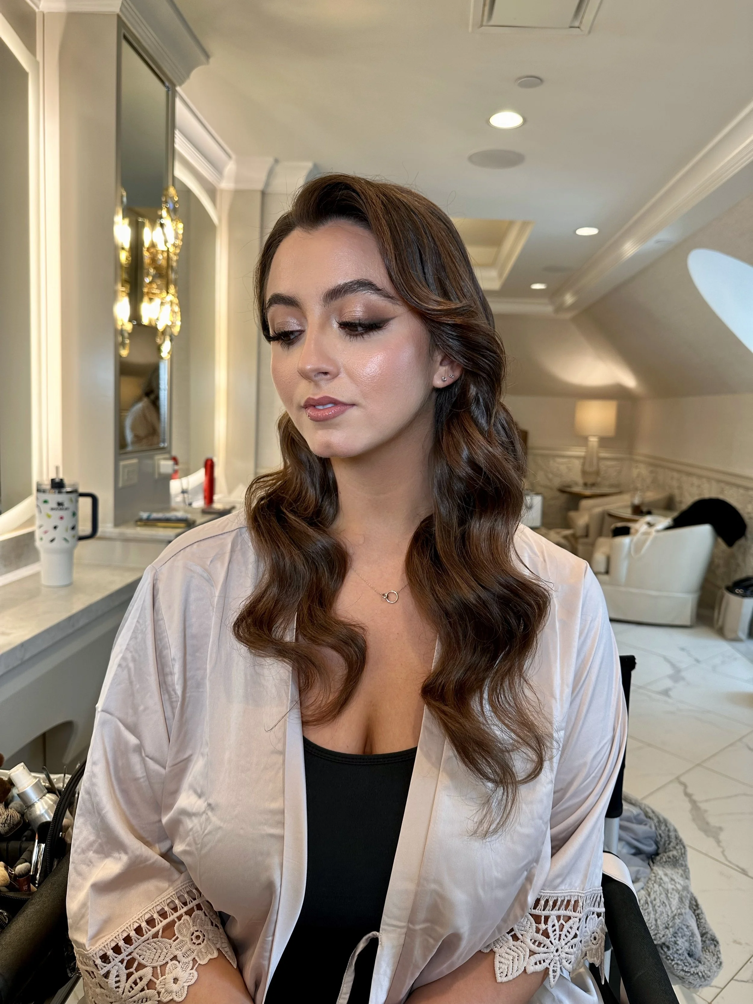 A woman with styled brown hair and makeup sitting in a room with beige walls, mirrors, and elegant lighting, possibly in a salon or dressing room.