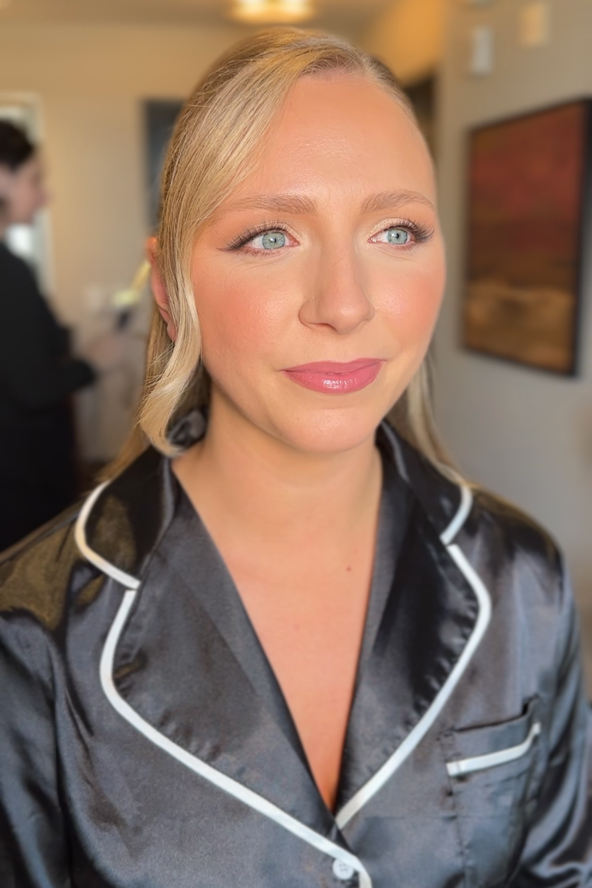 A woman with blonde hair and blue eyes wearing satin pajamas with white piping, standing in a room with blurred background, possibly a salon or dressing area.