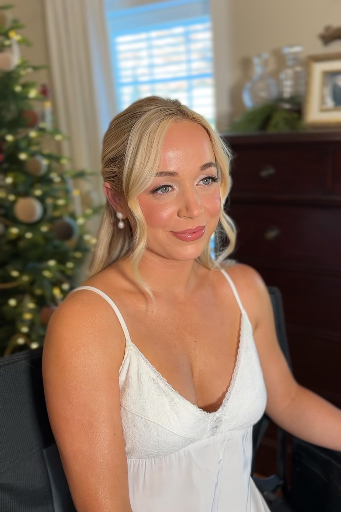 A woman with blonde hair wearing pearl earrings and a white spaghetti strap dress sitting indoors near a Christmas tree.