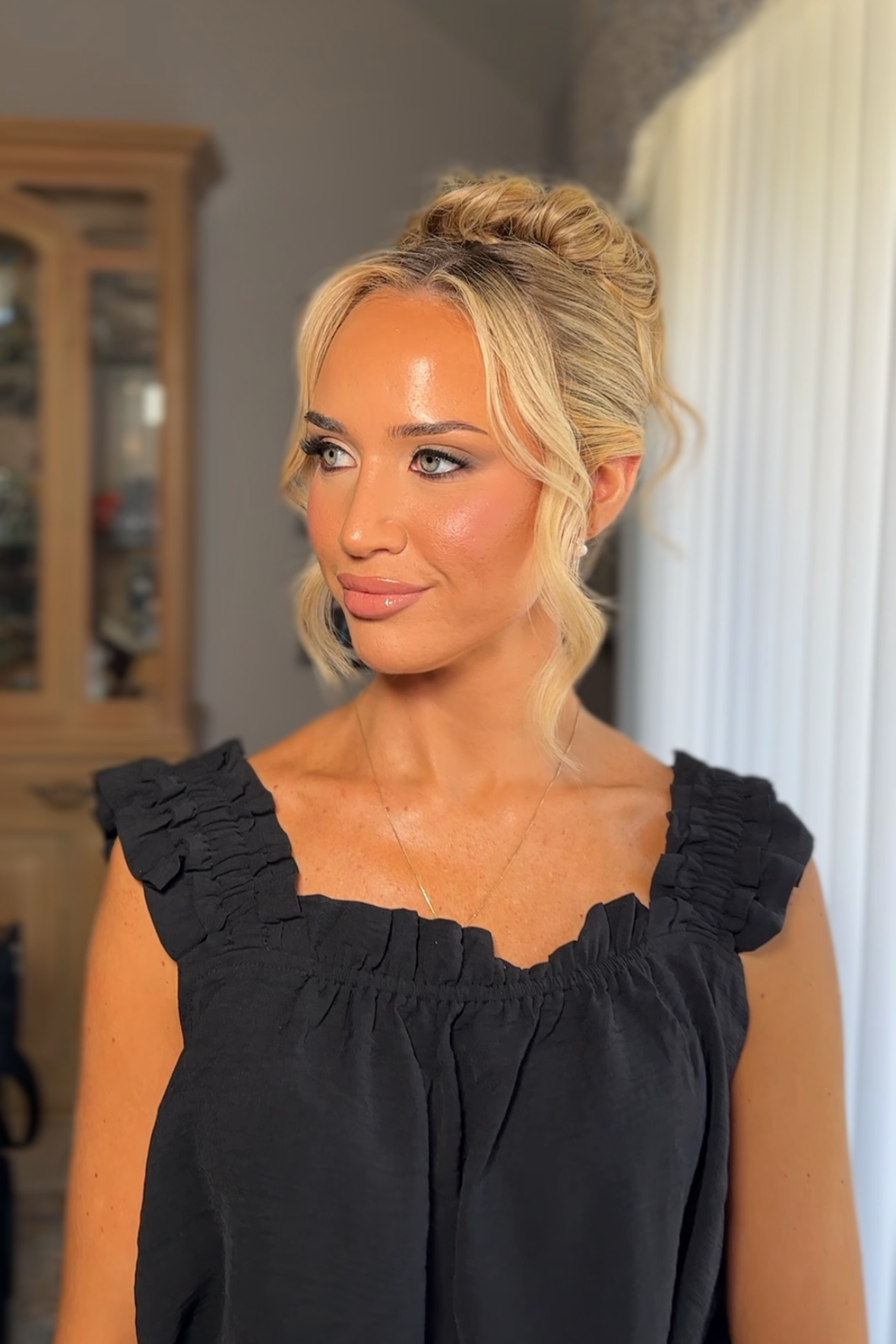 Portrait of a woman with styled blonde hair, wearing a black ruffled top, indoors near window with vertical blinds.