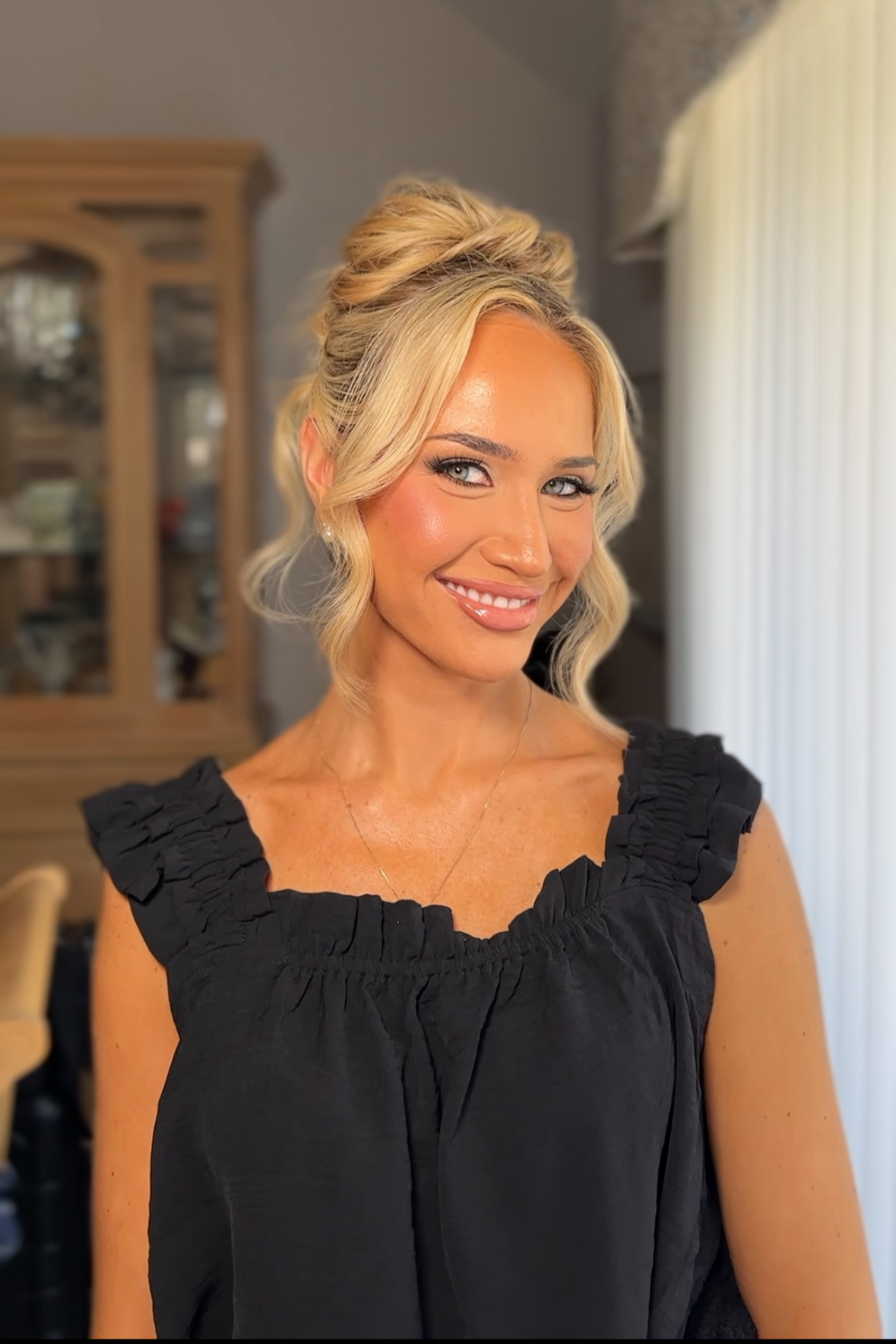 A woman with blonde hair styled in loose waves and an updo, smiling, wearing a black sleeveless top with ruffled shoulders, standing indoors in front of a window with blinds.