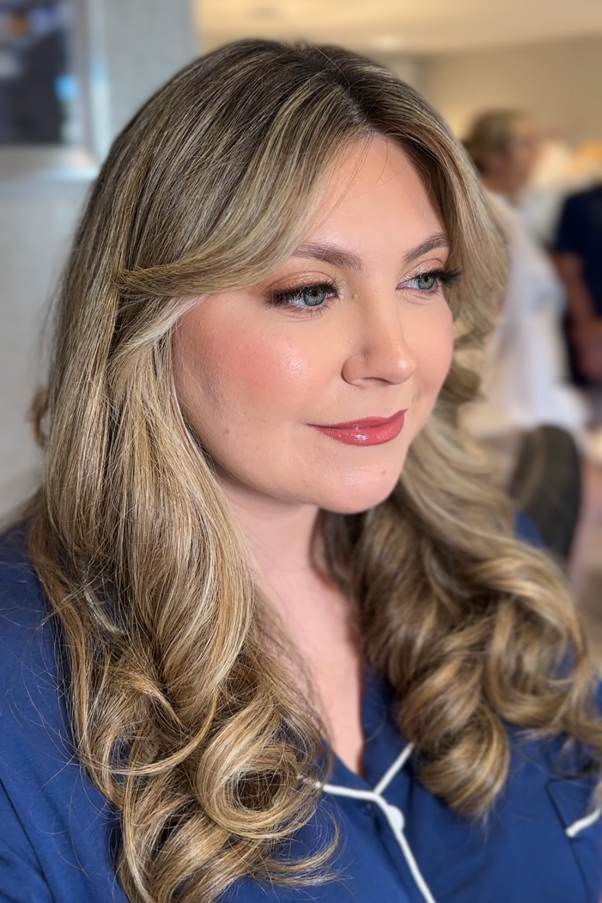 Close-up of a woman with styled blonde curly hair, makeup, and a navy blue shirt, sitting indoors with blurred people in the background.