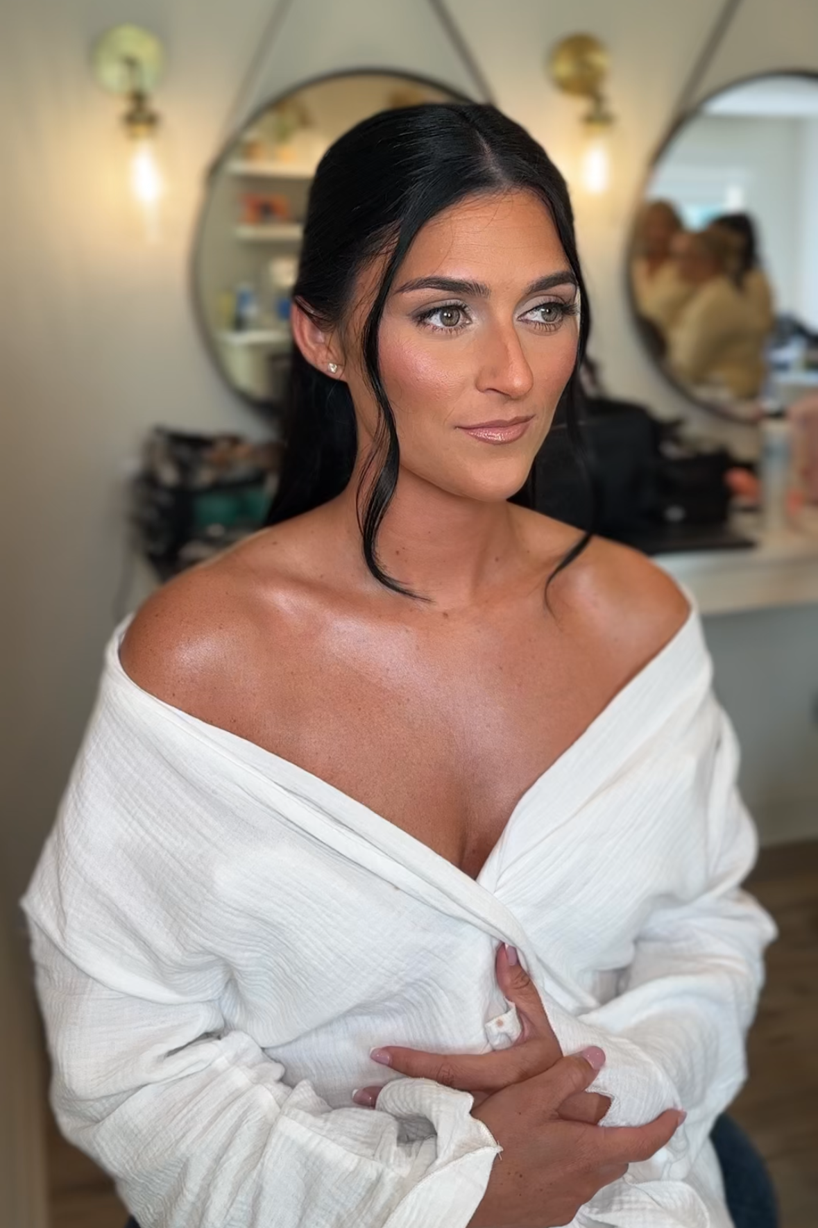 A woman with dark hair styled in loose waves, wearing makeup, and an off-shoulder white top, sits in a makeup chair in a salon or backstage setting with mirrors and shelves in the background.
