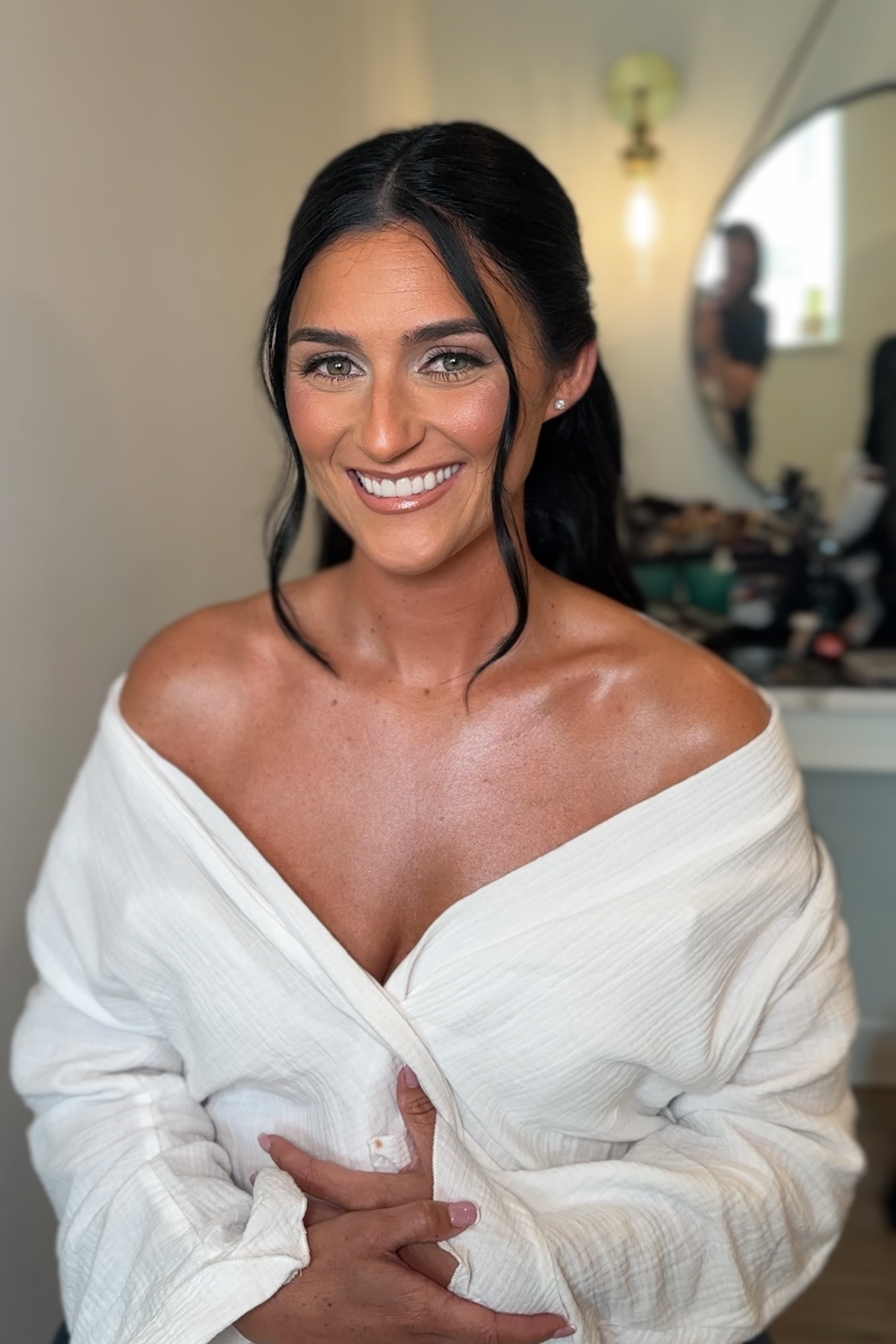 A smiling woman with dark hair in loose waves, wearing an off-the-shoulder white top, in a room with a large mirror and makeup items in the background.