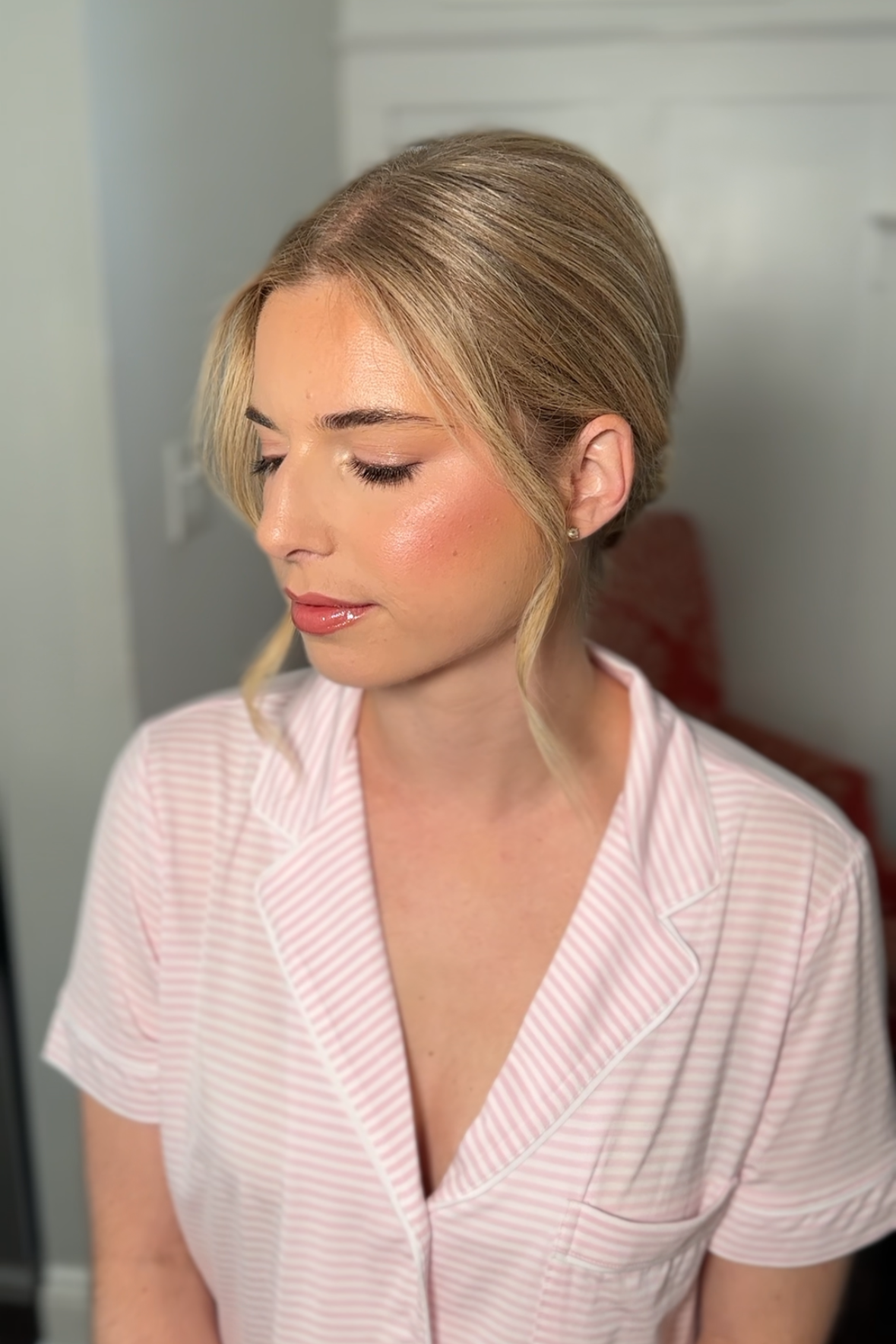 A woman with blonde hair styled in an elegant updo, wearing pink striped pajamas, looking down with her eyes closed, in a softly lit room.