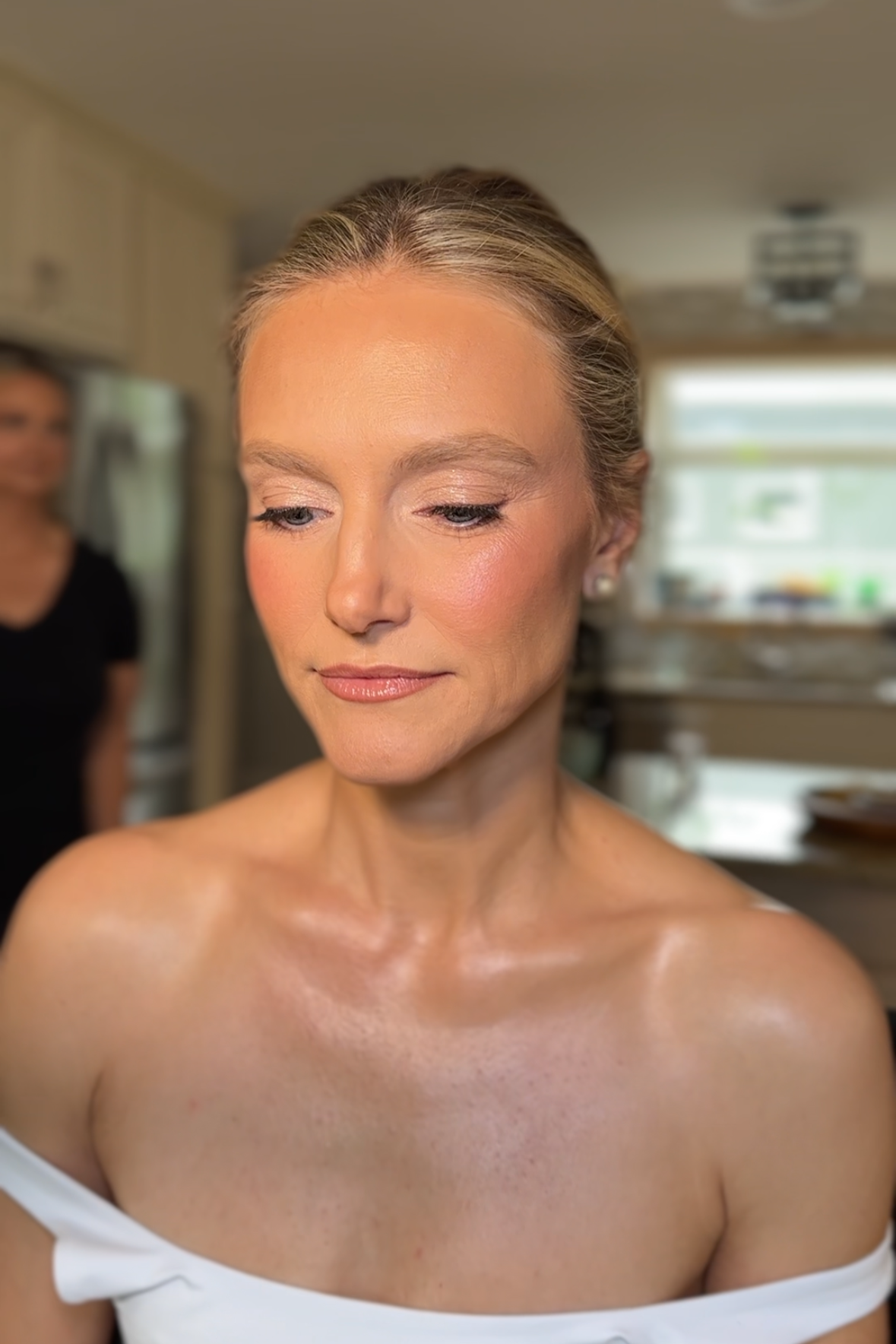 A woman with blonde hair styled in an updo, wearing makeup with pink lipstick and eye makeup, is looking down with a serene expression. She is in a kitchen, with a person in black standing in the background.