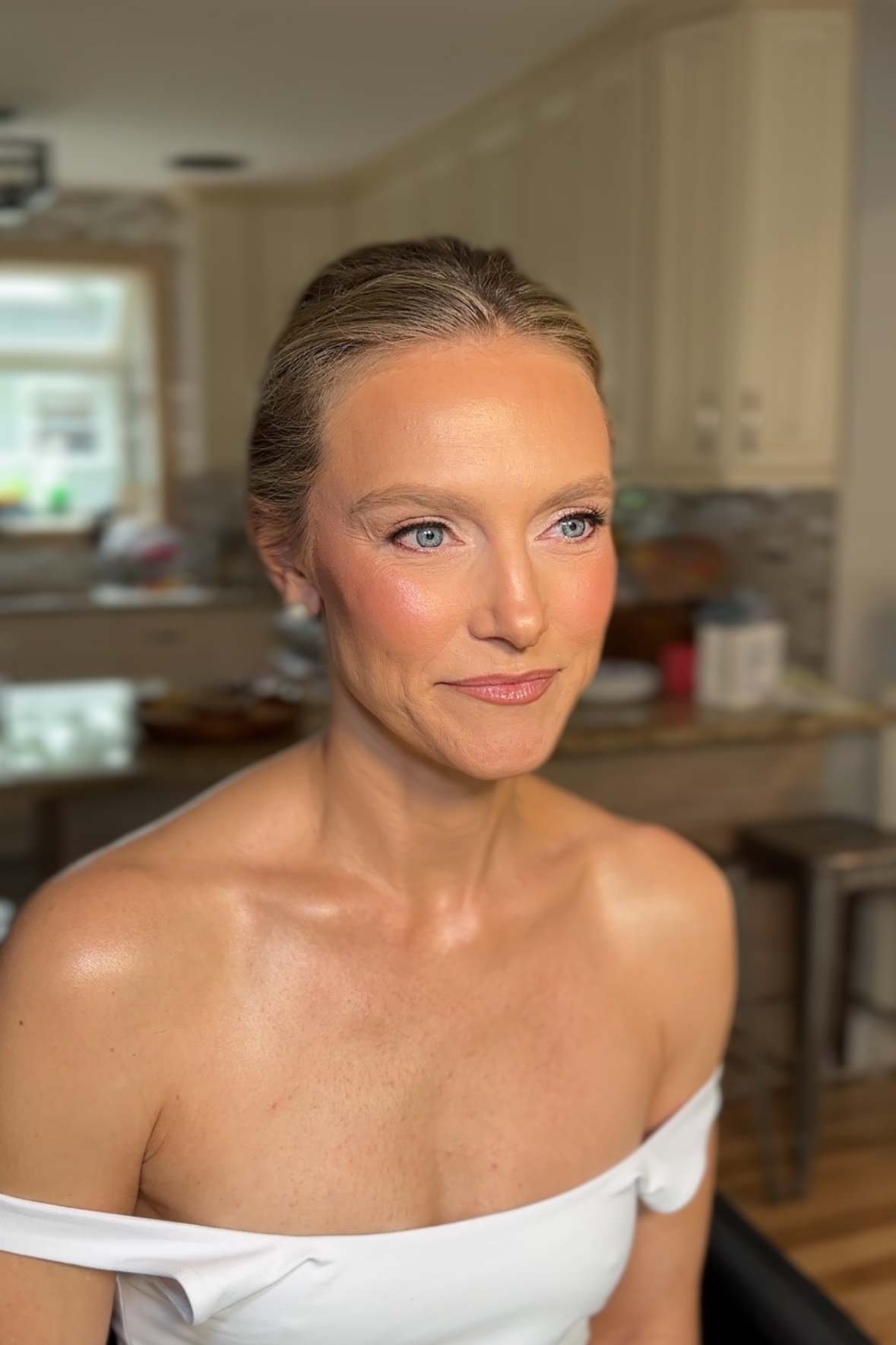 Close-up of a woman with light skin, blue eyes, and light brown hair styled back, wearing a white off-the-shoulder top, smiling softly, in a kitchen with cream cabinets and a window in the background.