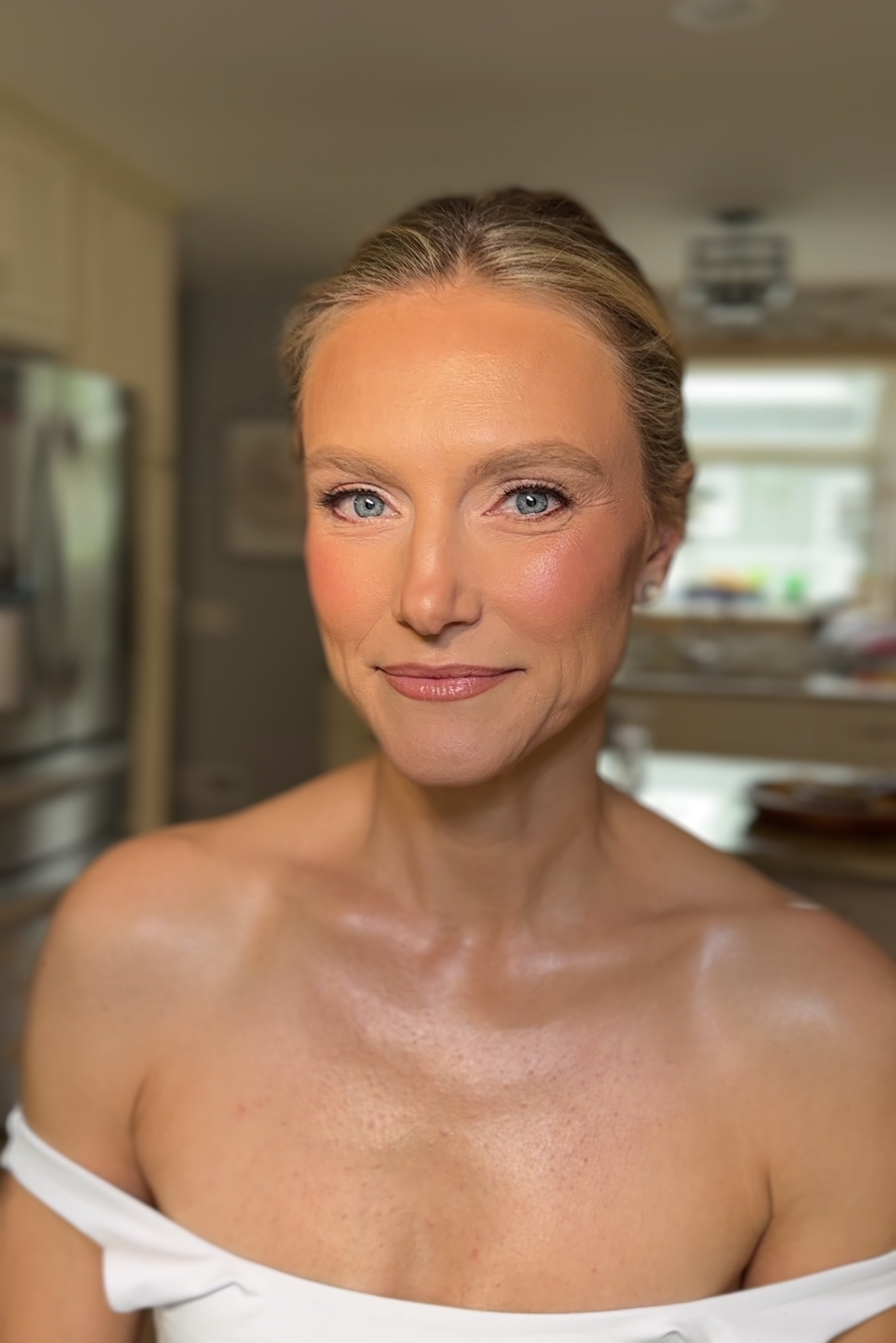 A close-up portrait of a woman with blonde hair, blue eyes, and light makeup smiling softly. She is wearing a white off-the-shoulder top and is indoors with a blurred kitchen background.