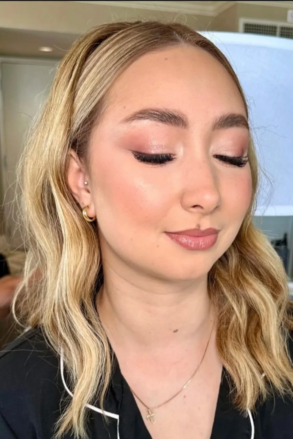 A young woman with blonde wavy hair, wearing makeup with pink eyeshadow and winged eyeliner, with closed eyes and a slight smile.