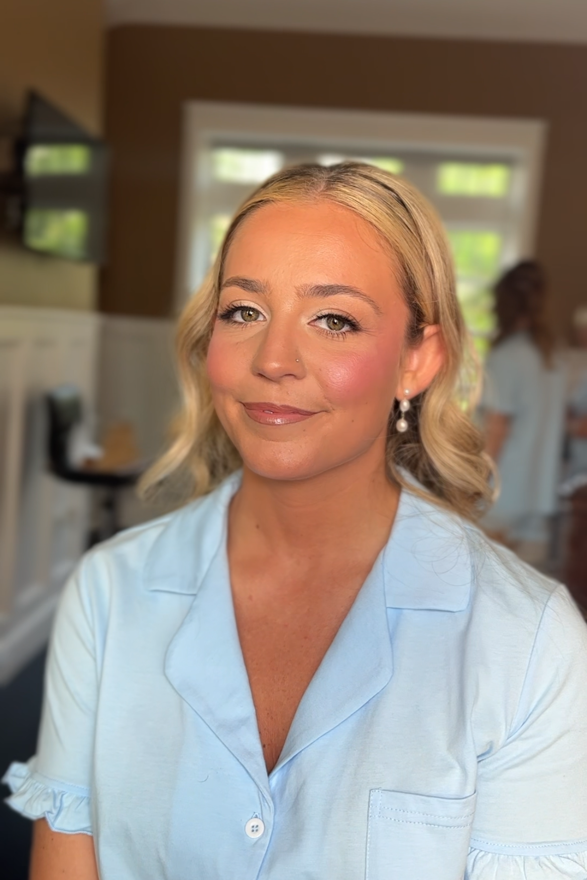 A woman with blonde hair, earrings, and a light blue shirt, smiling at the camera in a well-lit room with windows in the background.