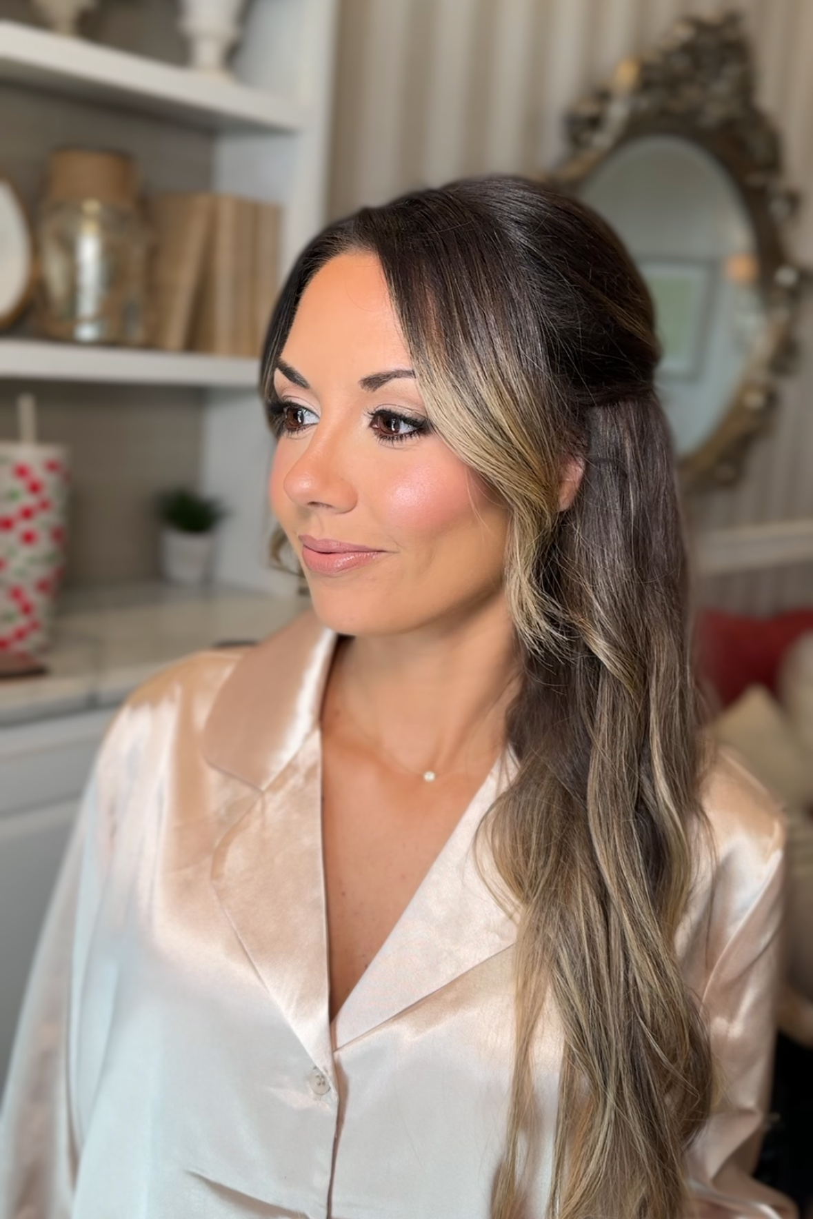 A woman with styled hair and makeup wearing a satin blouse in a cozy, decorated room.