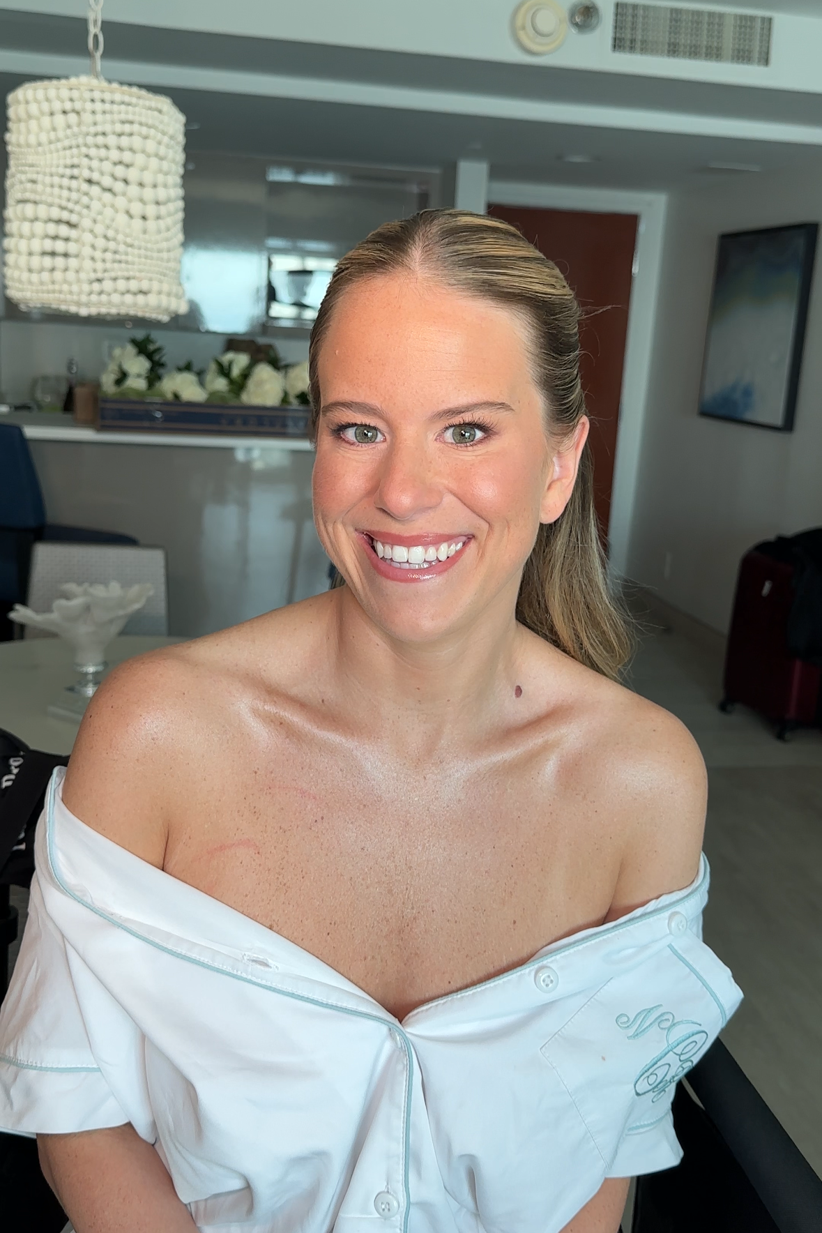 A woman with light brown hair in a half-up style, smiling, wearing an off-the-shoulder white shirt that is partially unbuttoned, in a bright, modern indoor space with flowers and furniture in the background.