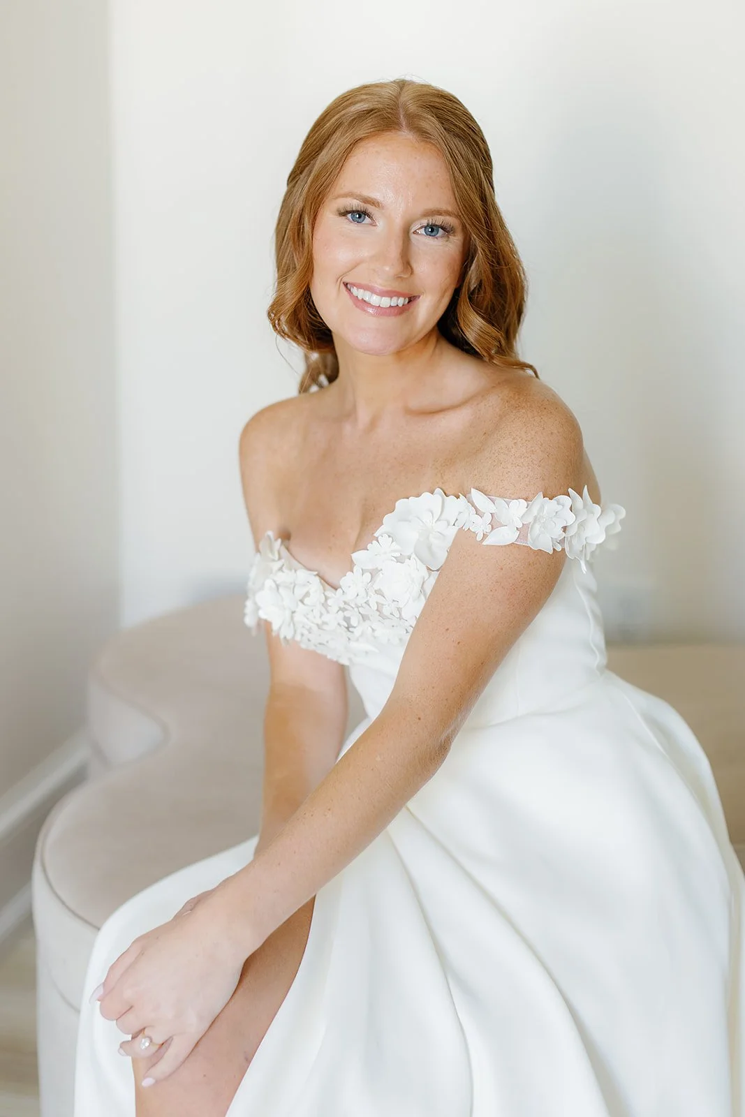 A smiling woman with red hair and blue eyes, wearing a white off-shoulder wedding dress with floral embellishments, sitting on a bench in a bright room.